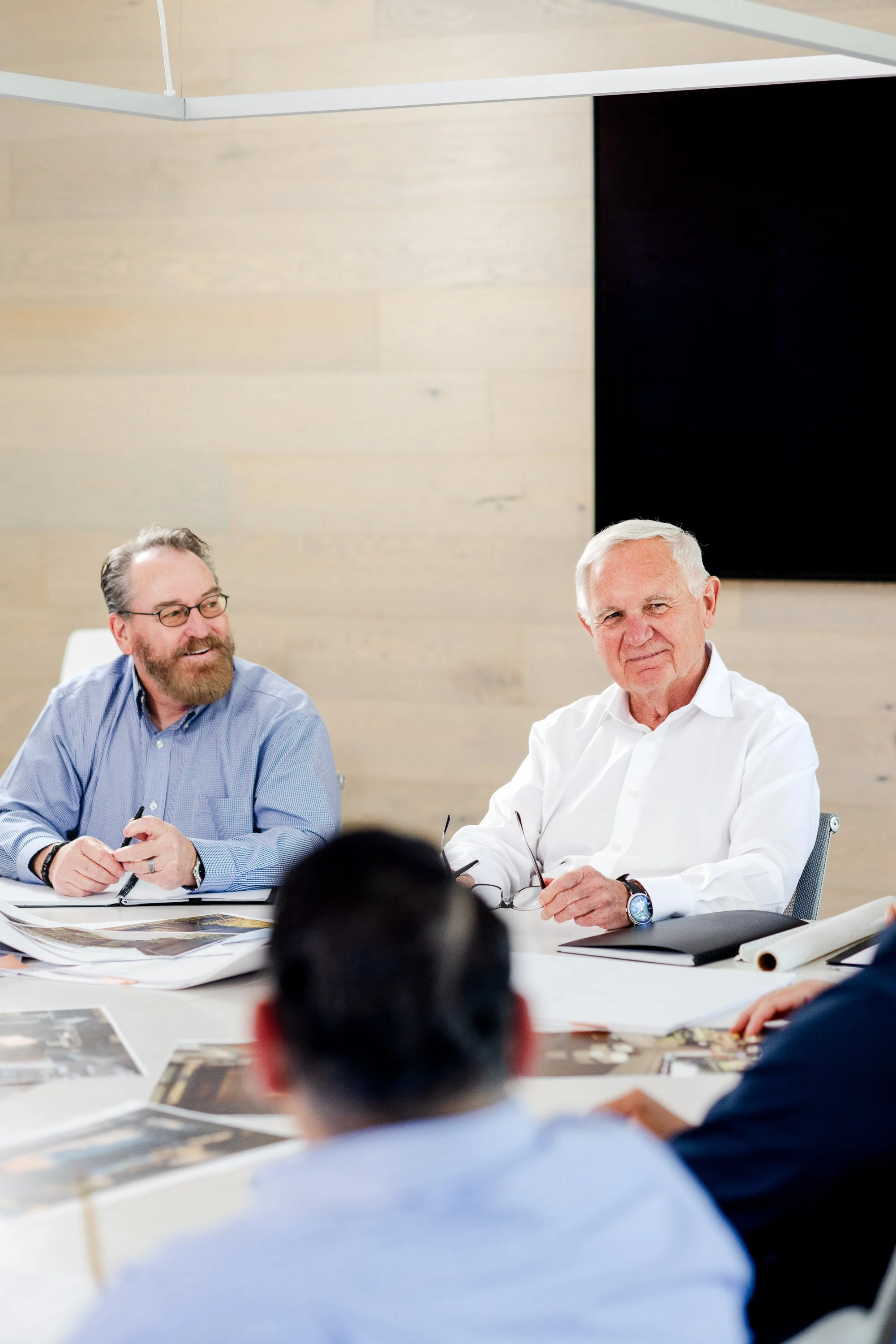 Two Robert Hidey Architects team members share a genuine smile during a group meeting, captured in a candid corporate lifestyle photograph.
