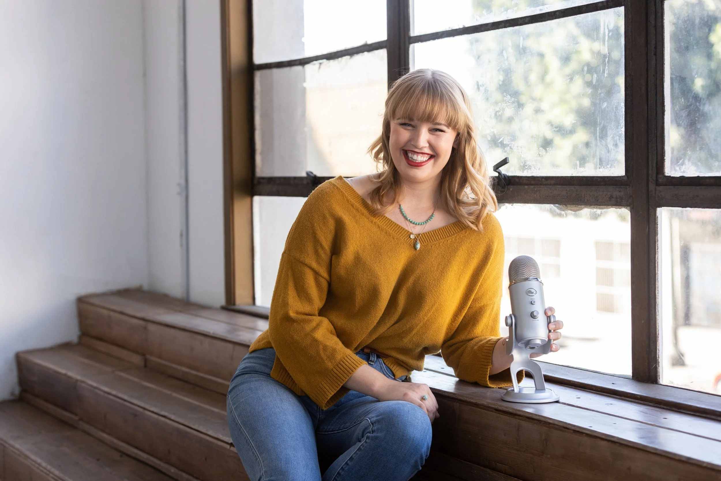 A blonde woman with a yellow sweater and jeans holds a Yeti podcast mic in this personal branding photo shoot for a women's business coach.