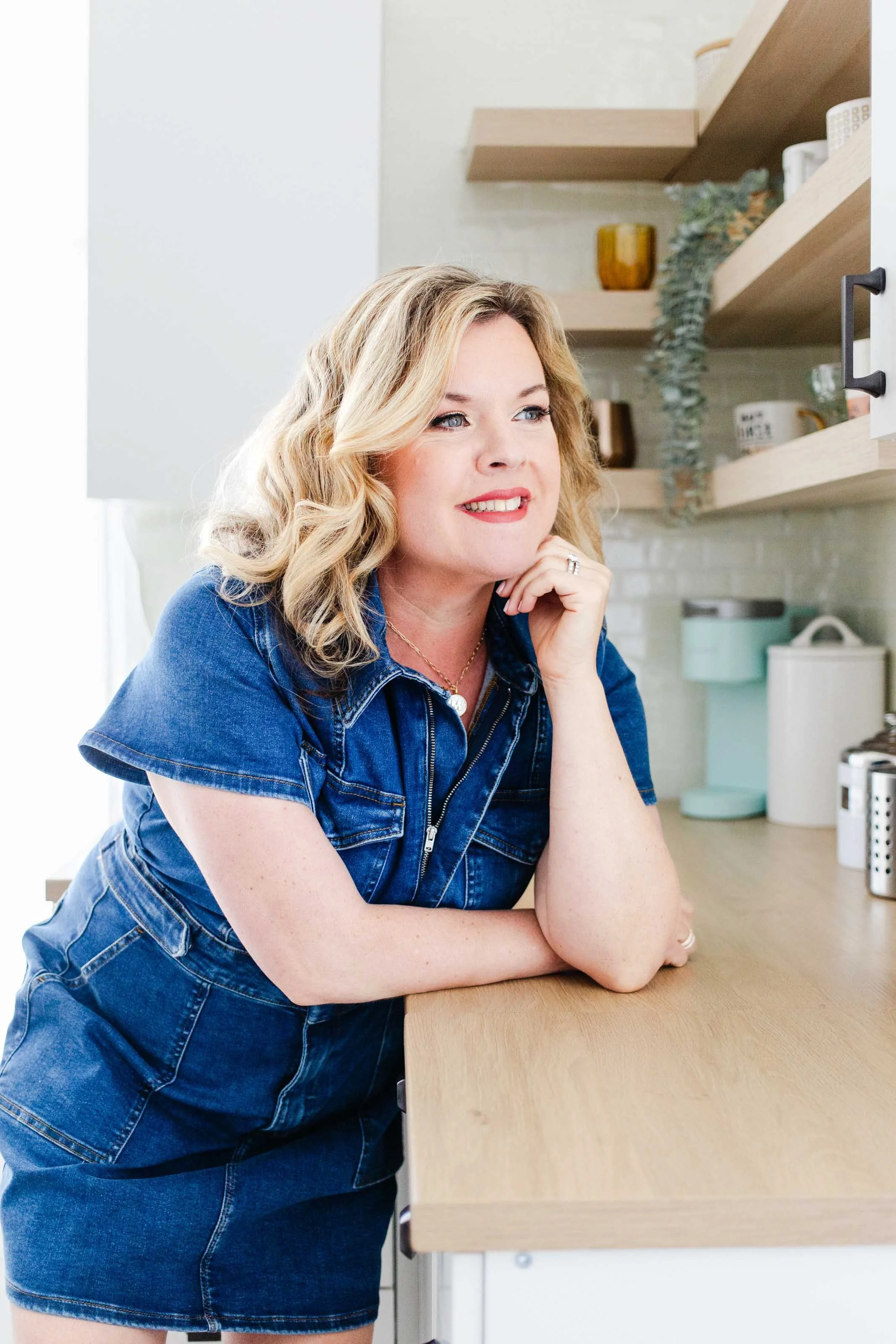 A blonde woman wearing a denim dress stands in a kitchen in this SF Bay Area brand photo shoot.