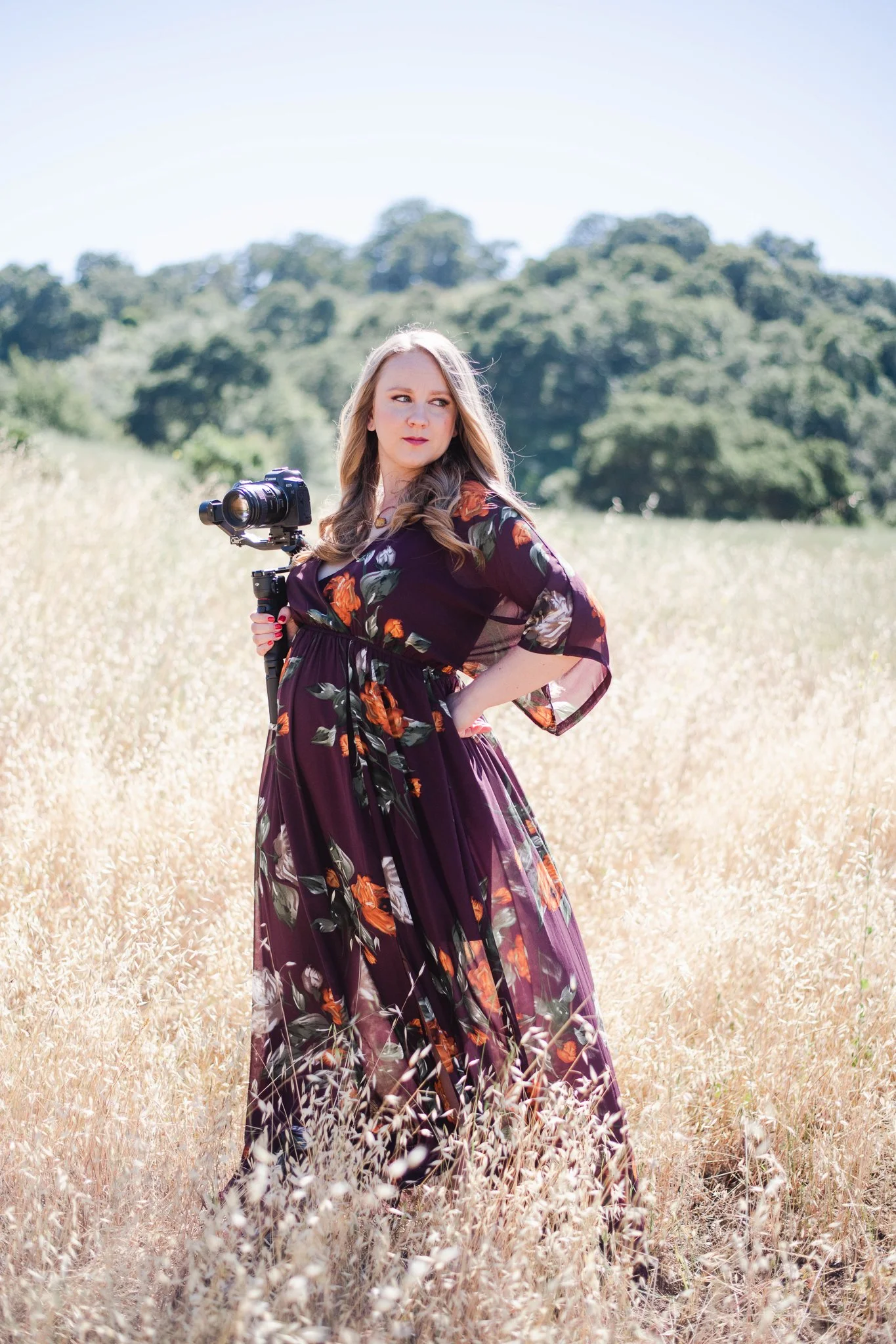 Brand photographer in a long flowy burgundy dress with flowers stands in a field with her camera.