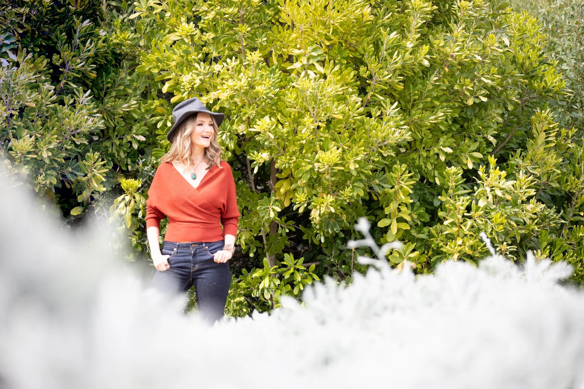 Outdoor fall brand photo of a woman in an orange sweater smiling on a sunlit path during a North Jersey brand shoot