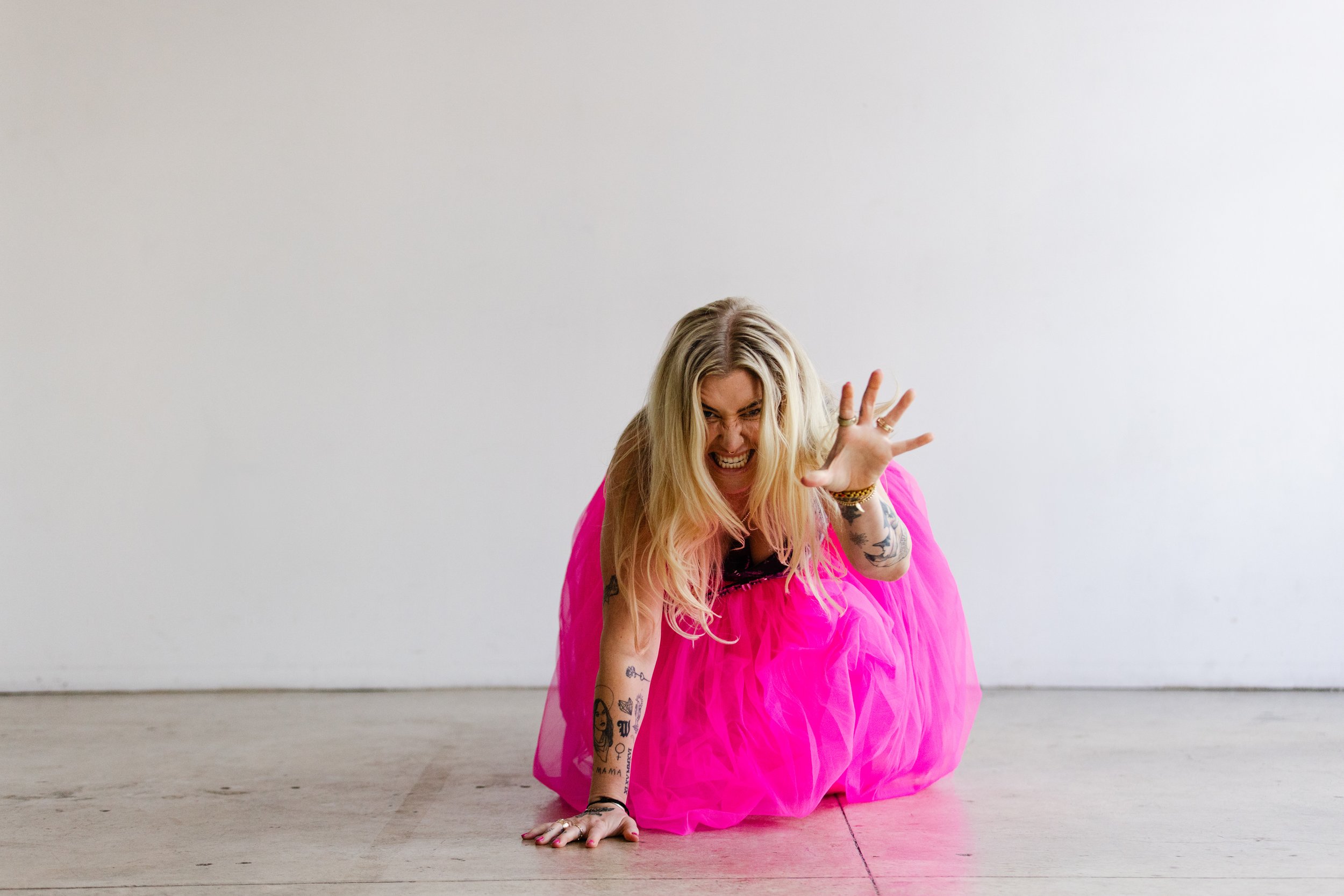 Dynamic branding shot of a woman in a hot pink tulle skirt crouching on the ground with a playful, fierce expression.