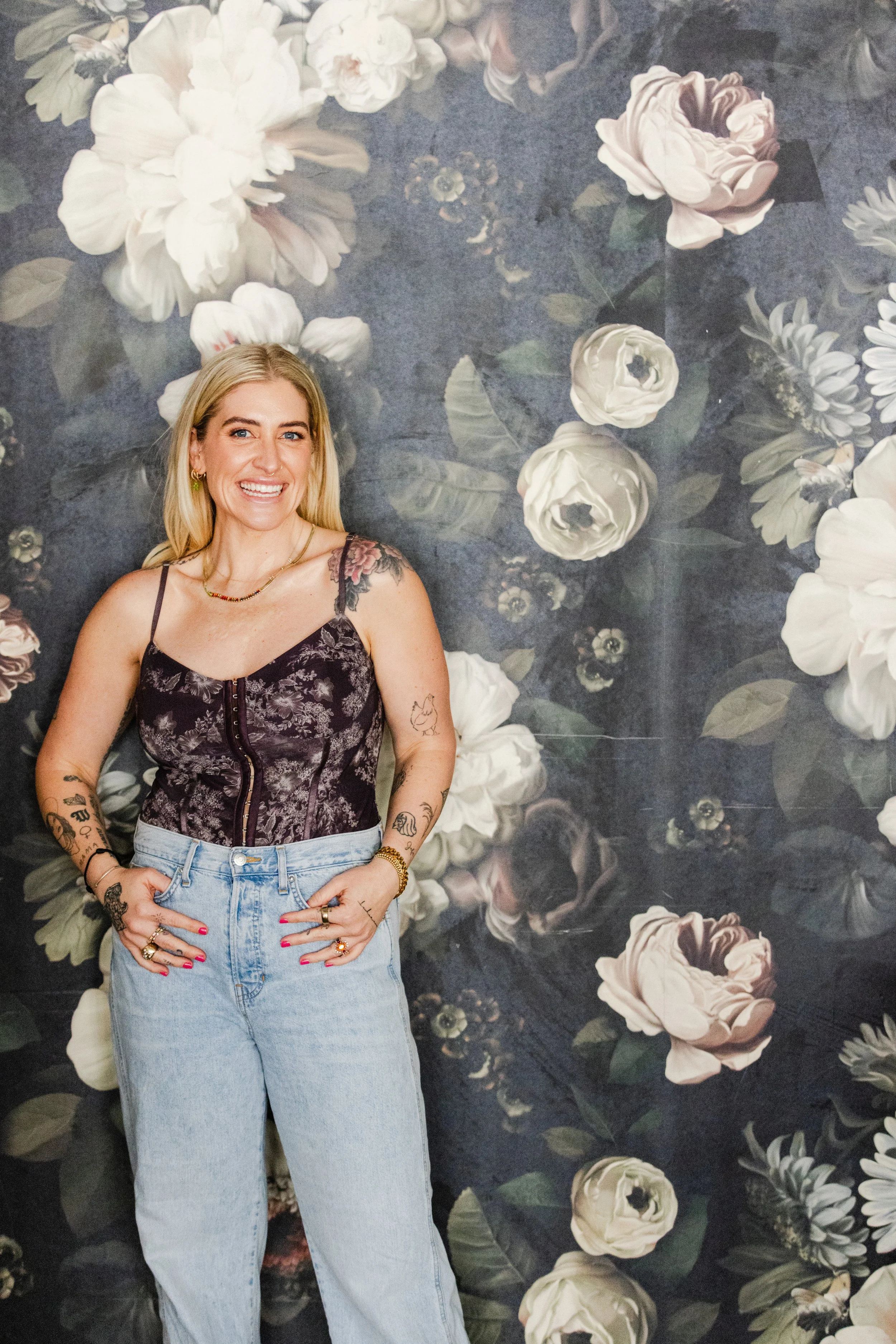 Portrait of a woman smiling in front of a dramatic floral wallpaper backdrop, wearing a dark corset top and light-wash jeans.