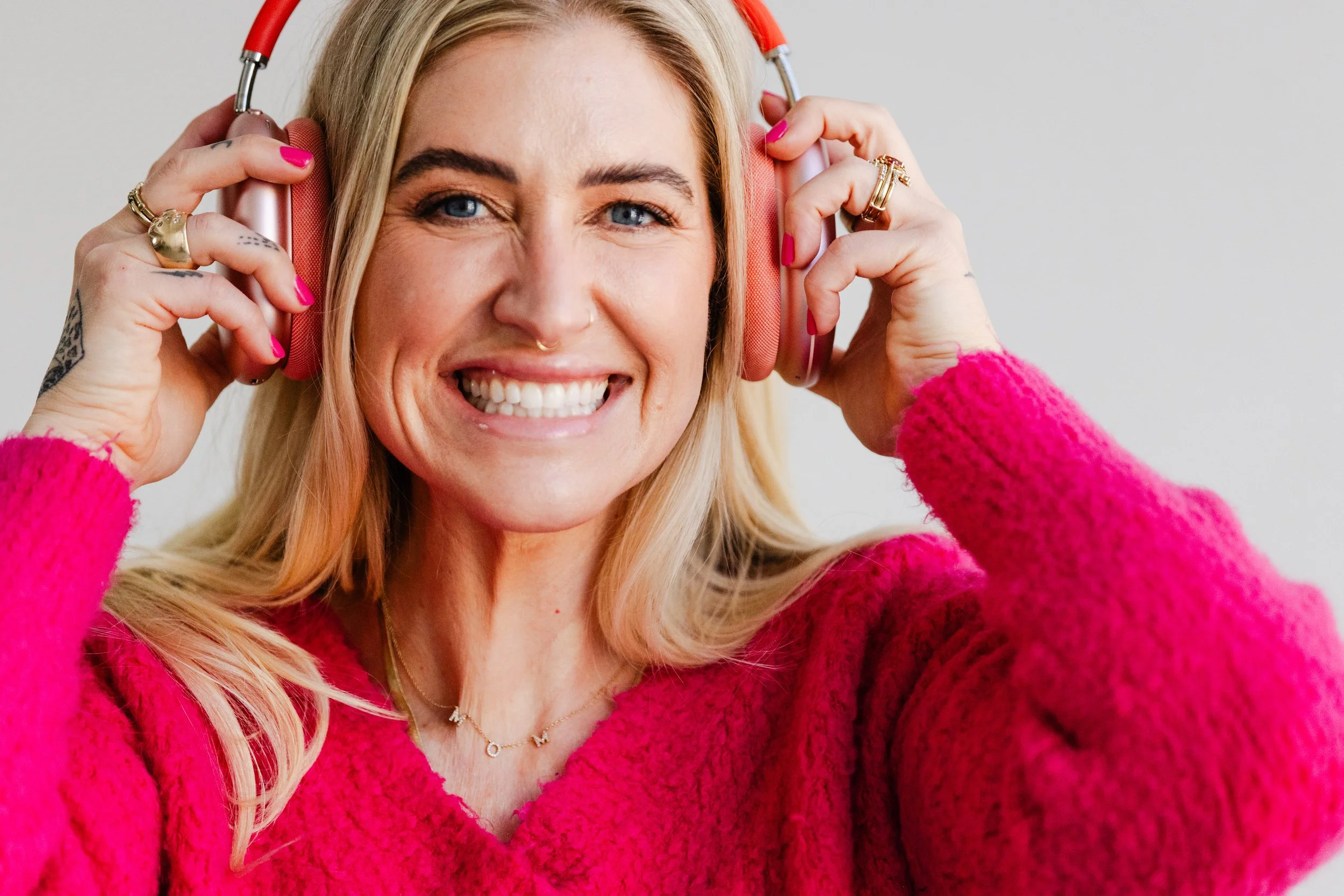 Close-up branding portrait of a smiling blonde woman in a bright pink sweater, holding coral-colored headphones to her ears.