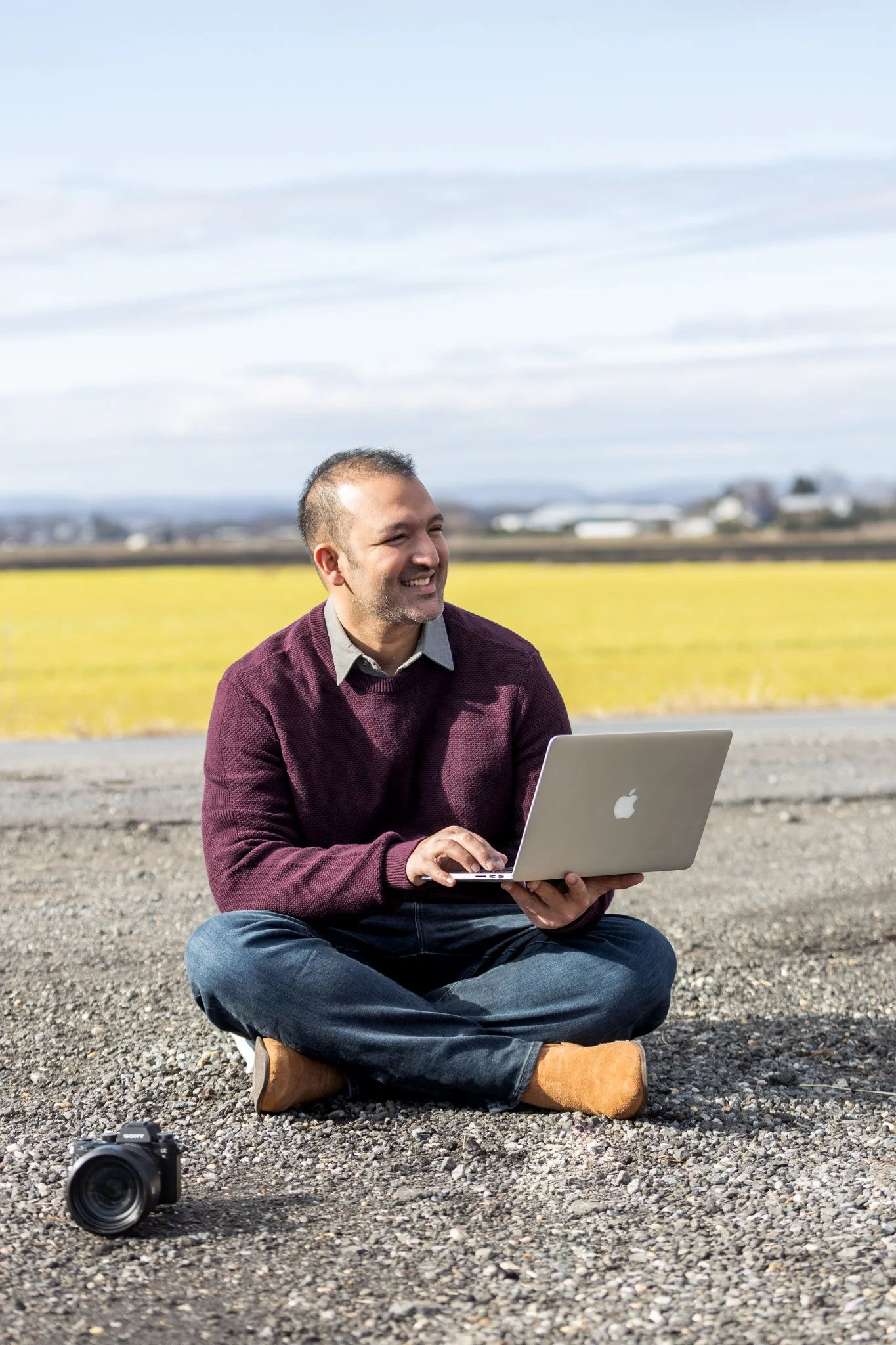 A man sits in a field with his laptop and camera for a NJ brand photoshoot.