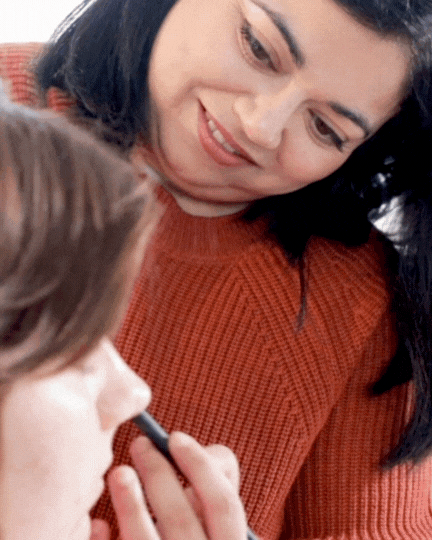 An animated image of a makeup artist with a burnt orange sweater applying eyeshadow to a woman with brunette hair.