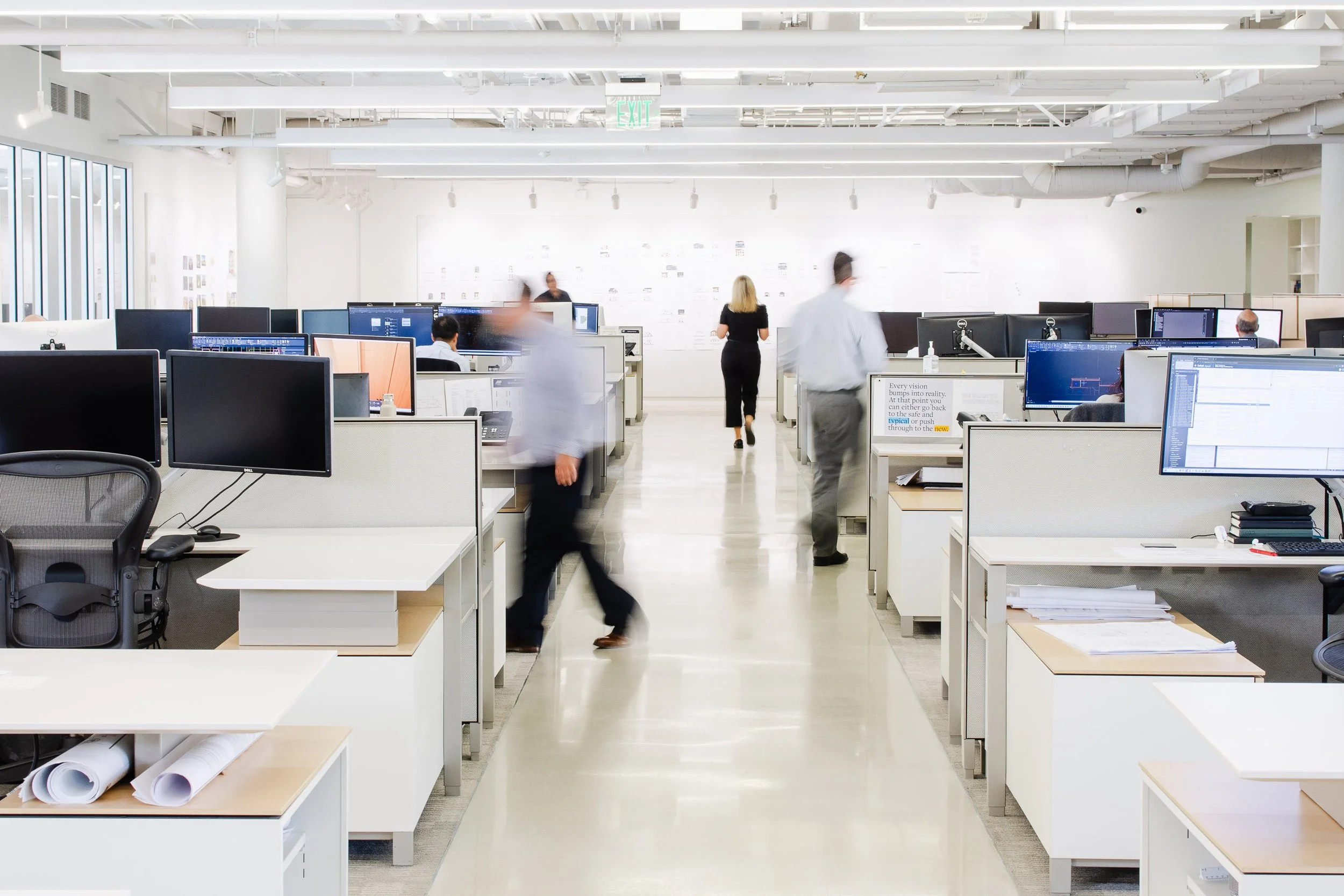 Wide environmental photograph of the Robert Hidey Architects office, showing an open workspace with employees moving between desks.