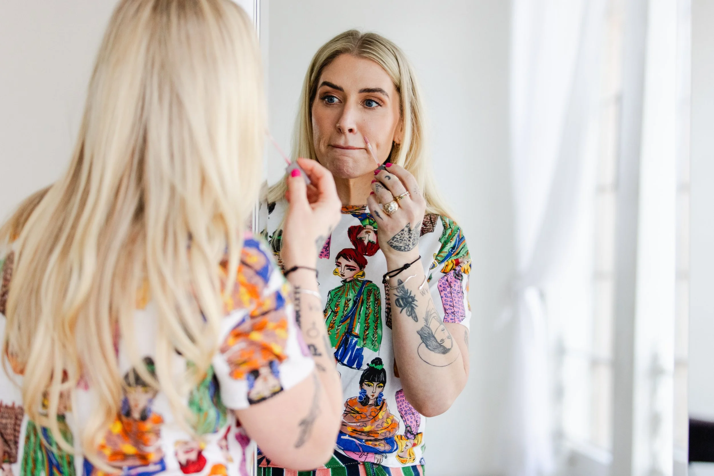 Candid photo of a woman applying lip gloss while looking into a mirror, wearing a colorful illustrated top and standing in a softly lit room.