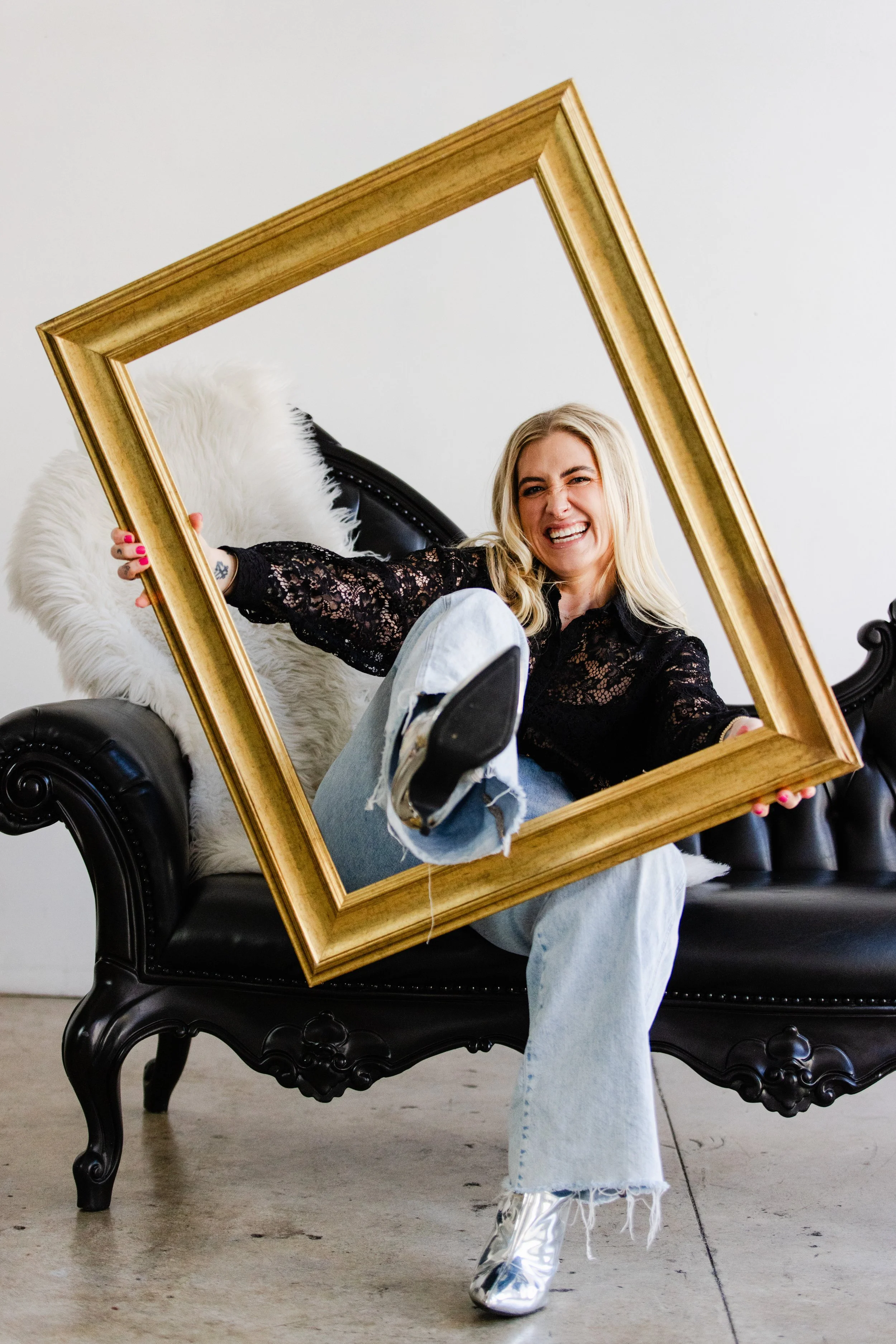 Los Angeles business coach smiling playfully while holding a gold frame around herself, seated on a black vintage couch in a personal brand photo shoot.