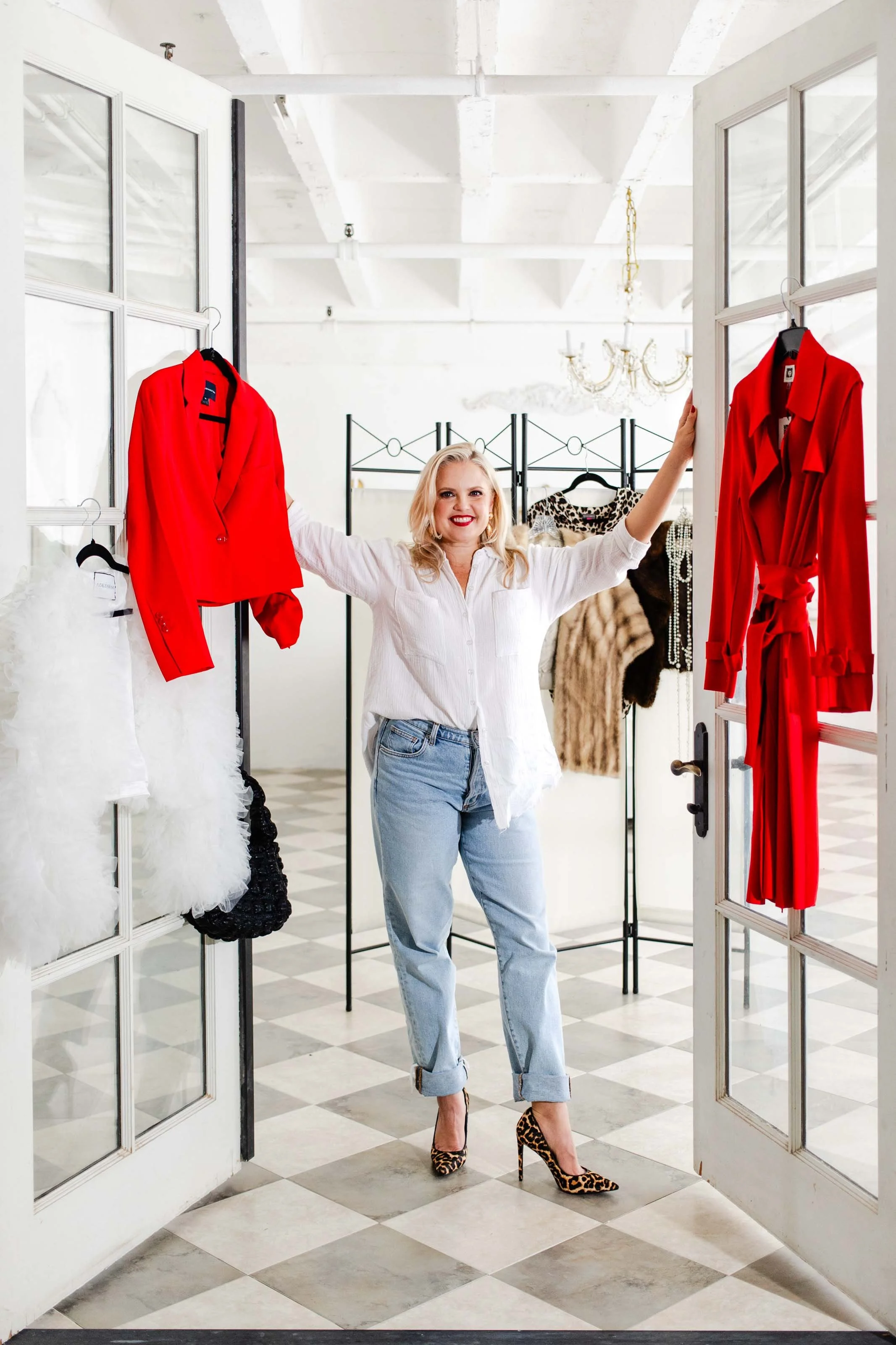 Blonde woman with red lipstick during a Los Angeles brand photo shoot opening double doors to a room with a wardrobe rack. She is dressed in a white button up top and light colored jeans. There are two red jackets hanging up amongst the background.