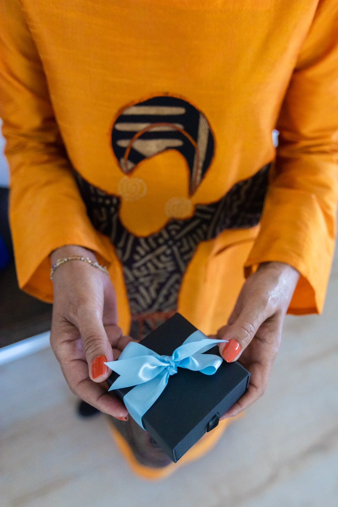A black woman in an orange dress holds a box with a blue ribbon during a California brand photography session with Bernadette Marciniak of Sol Haus Media