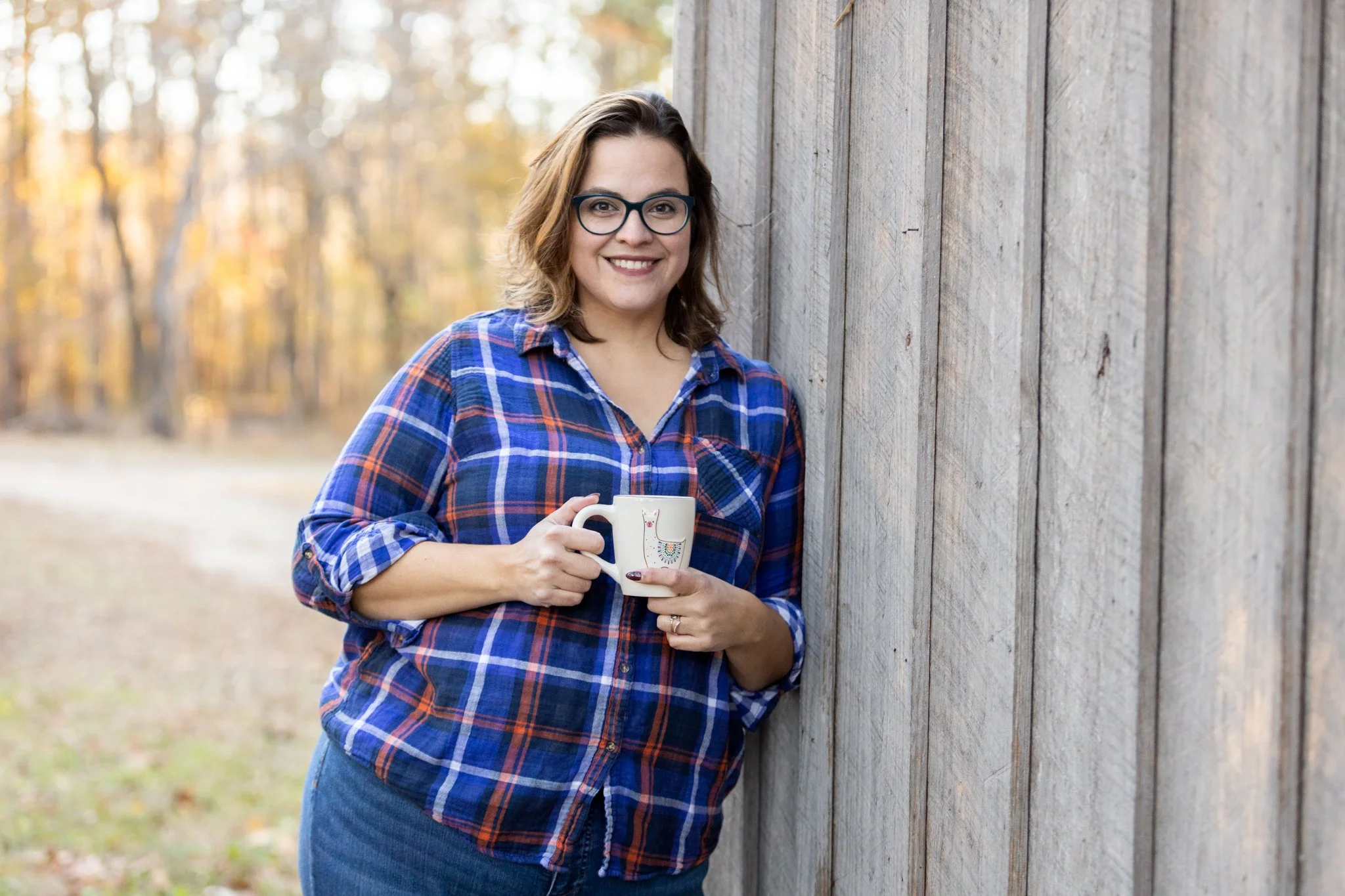 A brunette white woman in a blue plaid shirt leans up against a wooden barn with a white mug for her personal branding photo shoot.