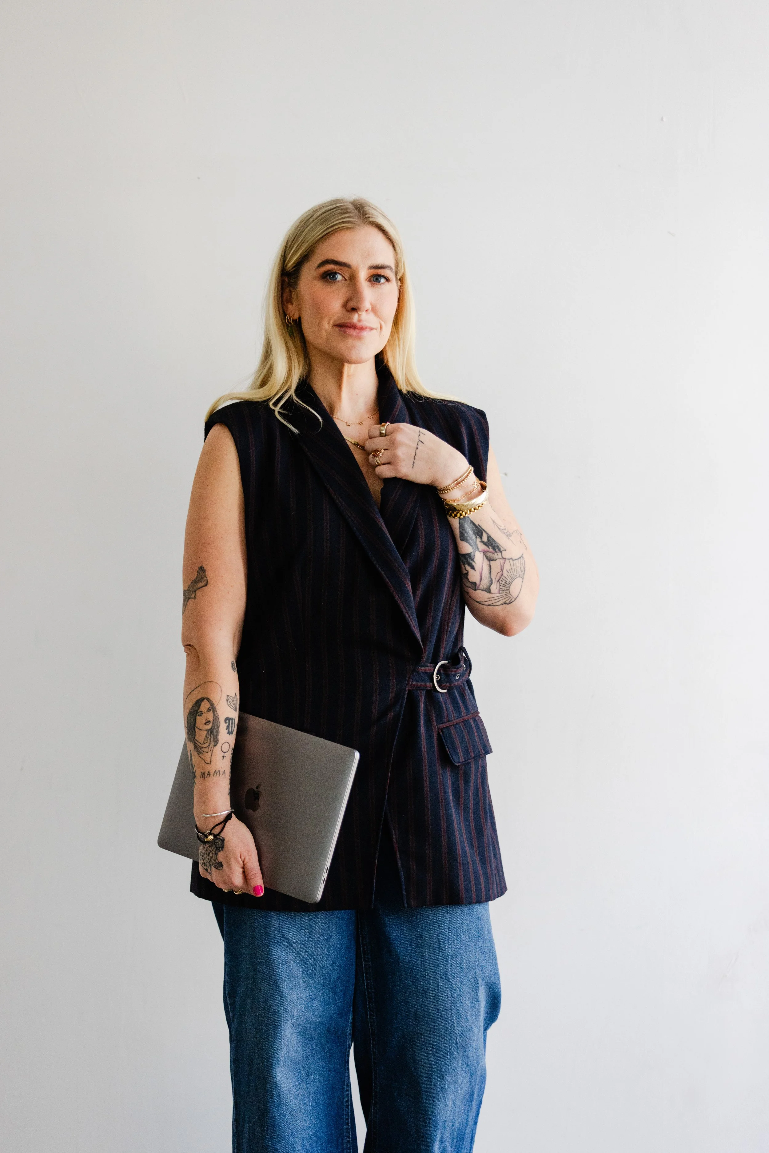 A business coach posing confidently with her laptop, showing off tattoos and a pinstripe vest in a modern brand photography session in Los Angeles.