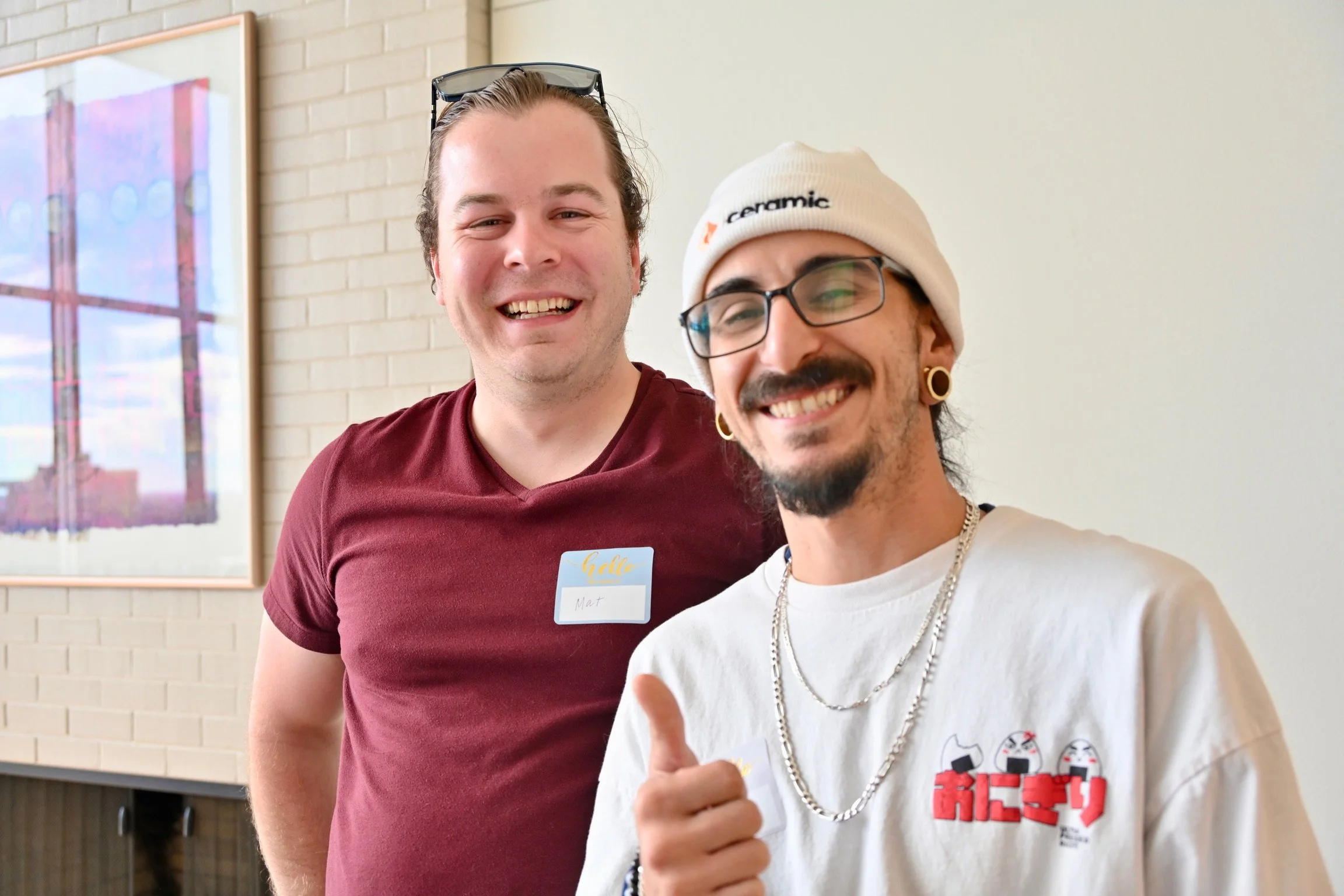 Two smiling men standing indoors, one wearing a maroon shirt and the other wearing a white shirt with Japanese characters, necklaces, a beige beanie, and glasses, giving a thumbs-up gesture.