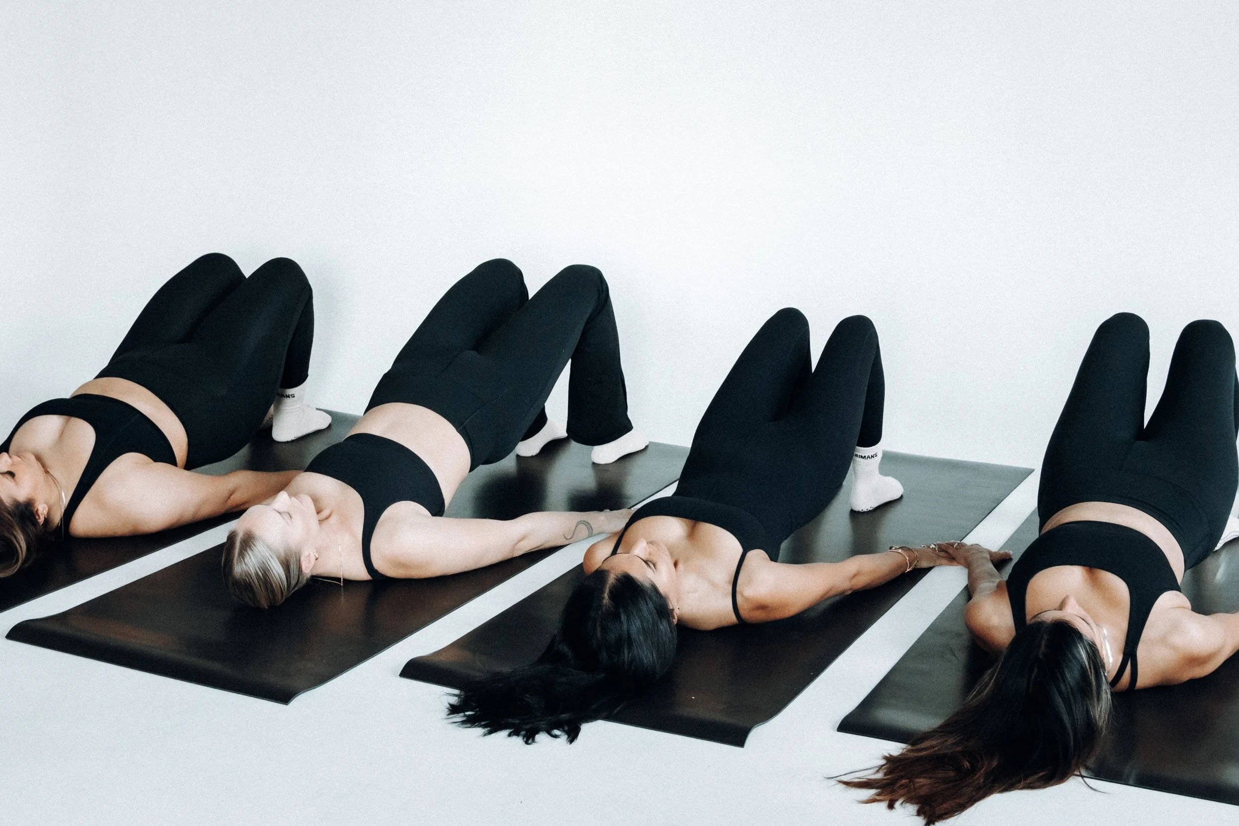 Three women lying on black yoga mats, practicing relaxation or meditation, with closed eyes, in a minimalist room.