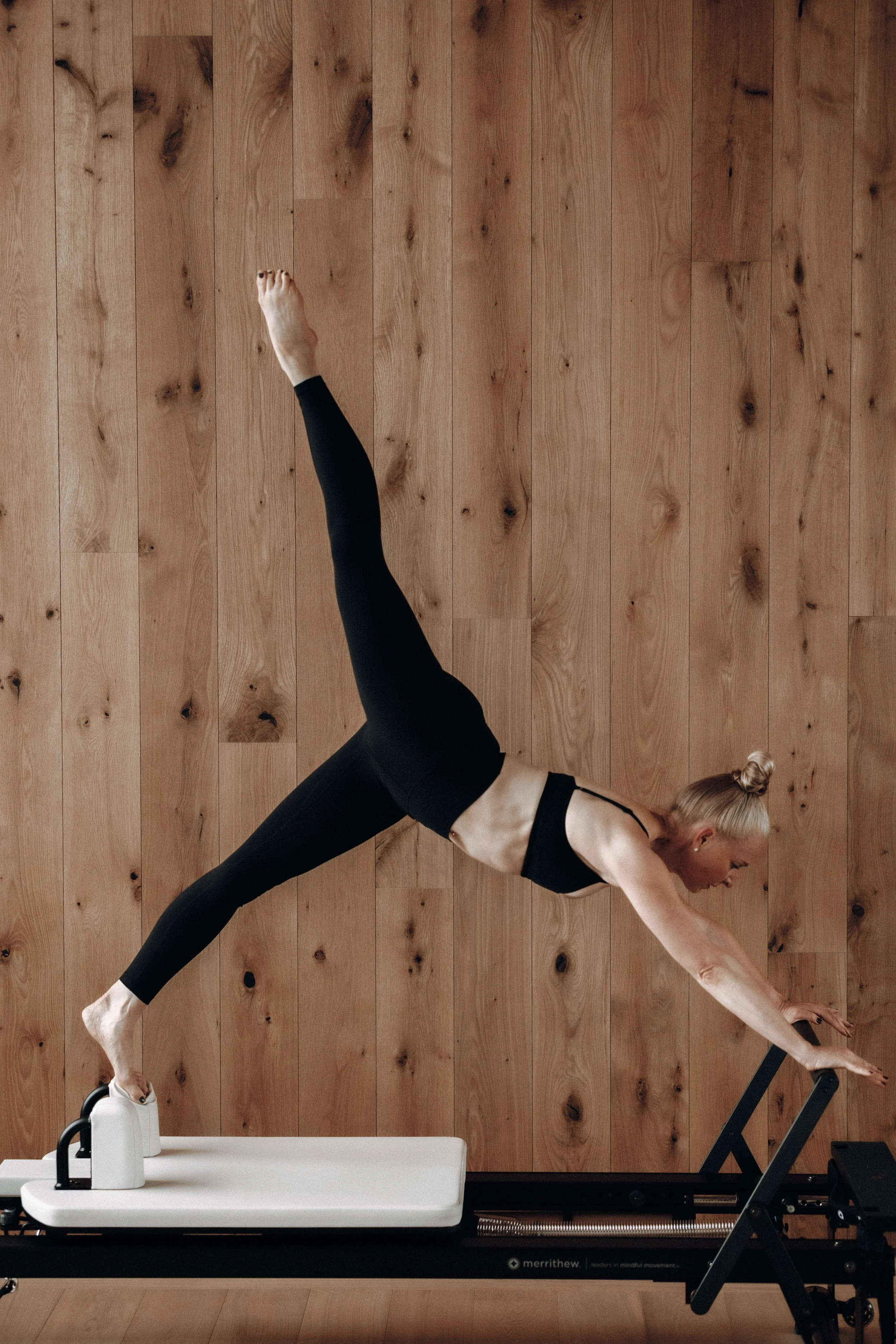 Woman in black athletic wear performing a plank exercise on a Pilates reformer machine against a wooden wall.