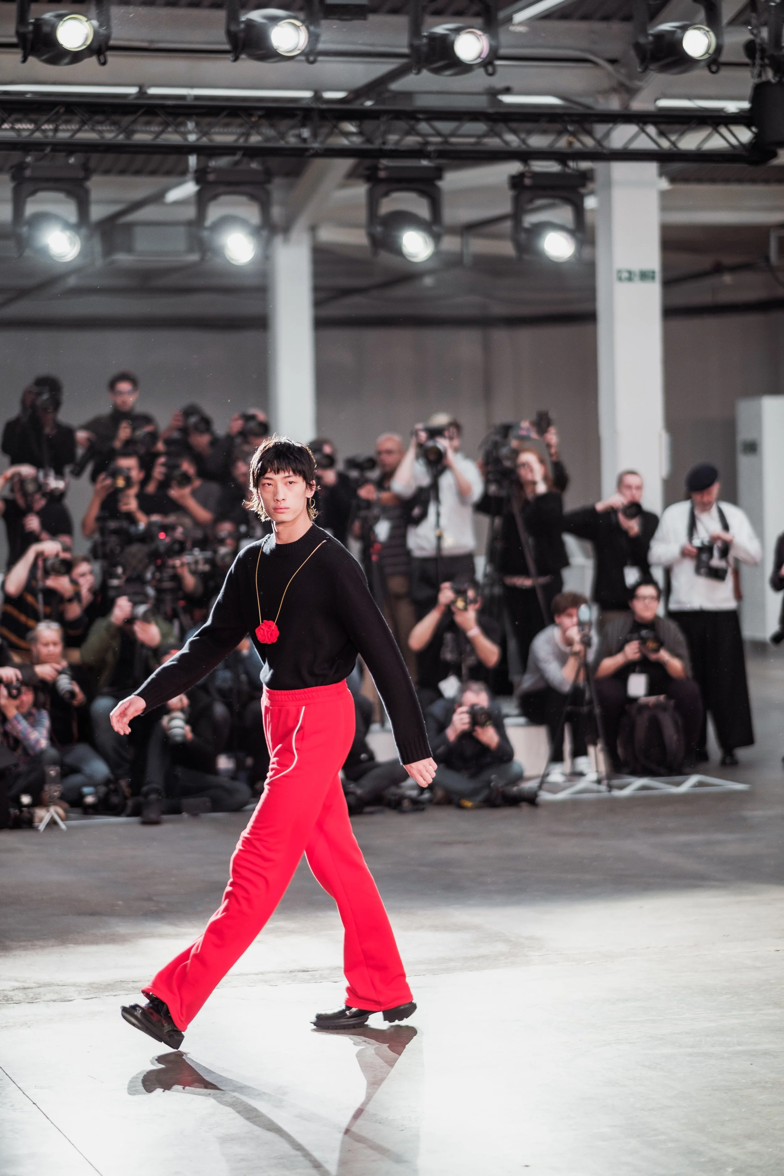 A fashion runway show with a model walking on the runway in front of a large group of photographers and spectators, under bright overhead lights.