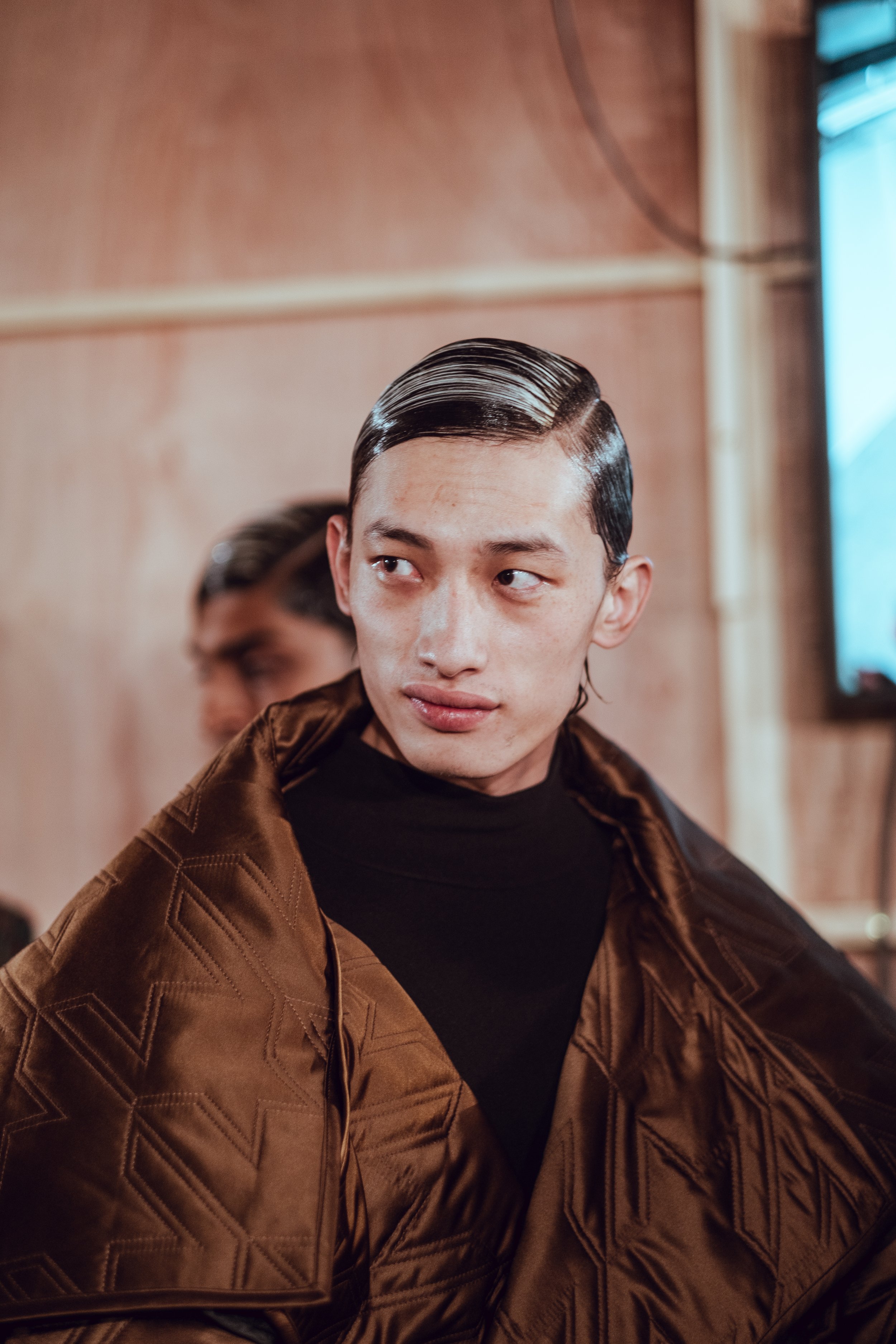 A young man with slicked-back black hair wearing a brown textured jacket and black shirt, looking to the side. He is indoors with a wooden wall and a window in the background.