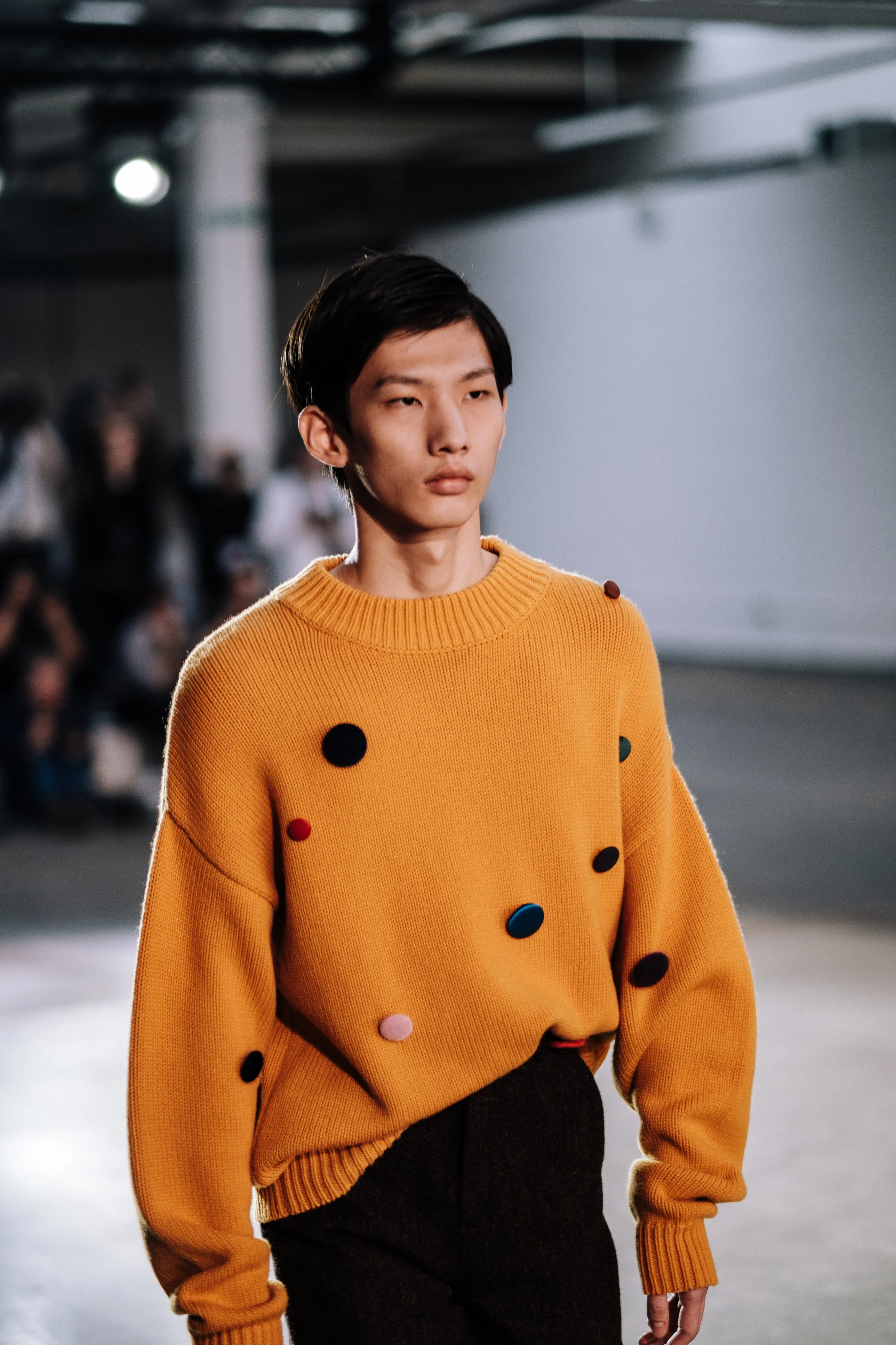 A young person walking on a runway wearing a yellow sweater decorated with colorful pom-poms, with a faint crowd in the background.