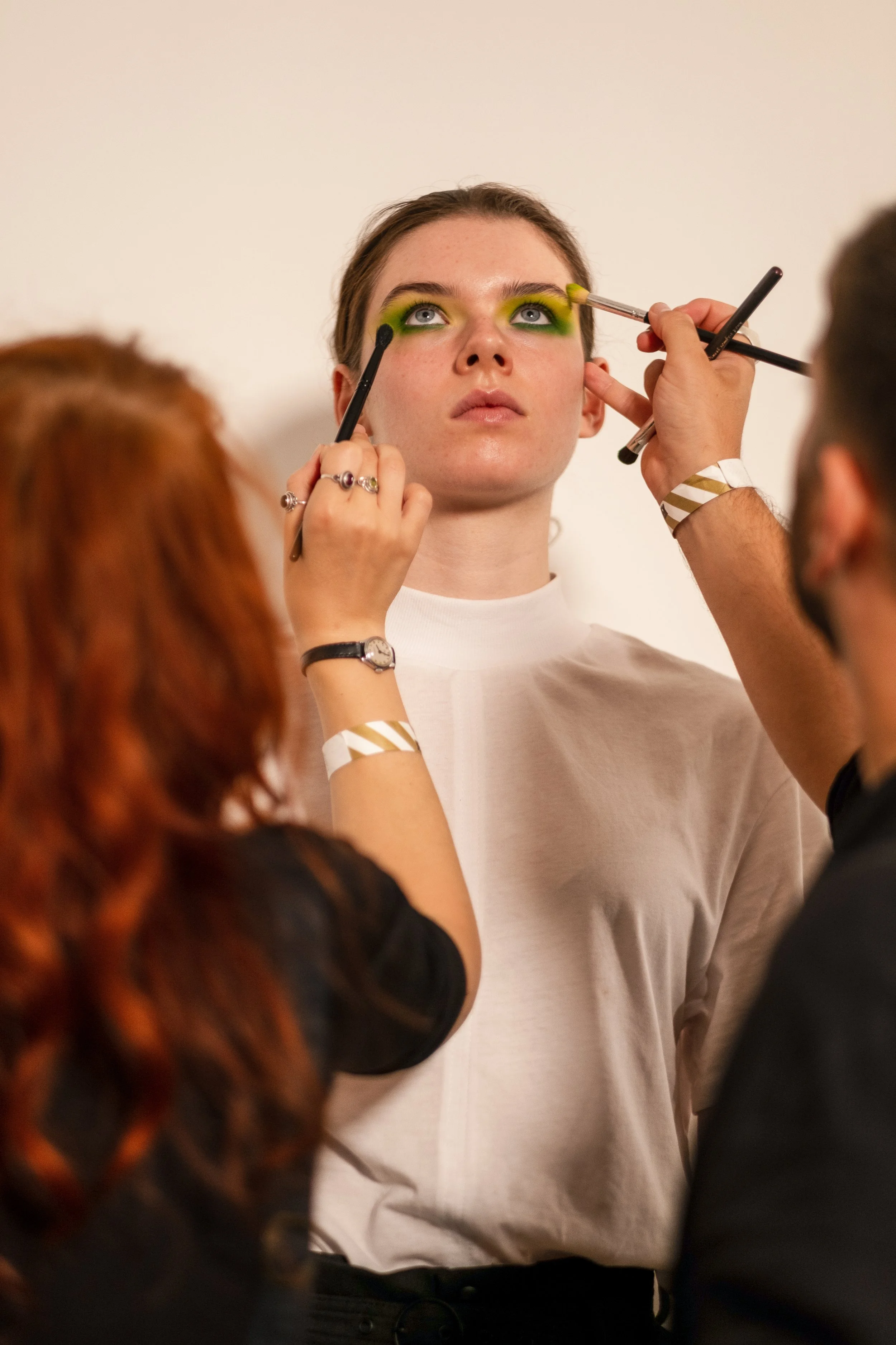 A young woman is having her makeup done by two artists, applying green eyeshadow around her eyes.