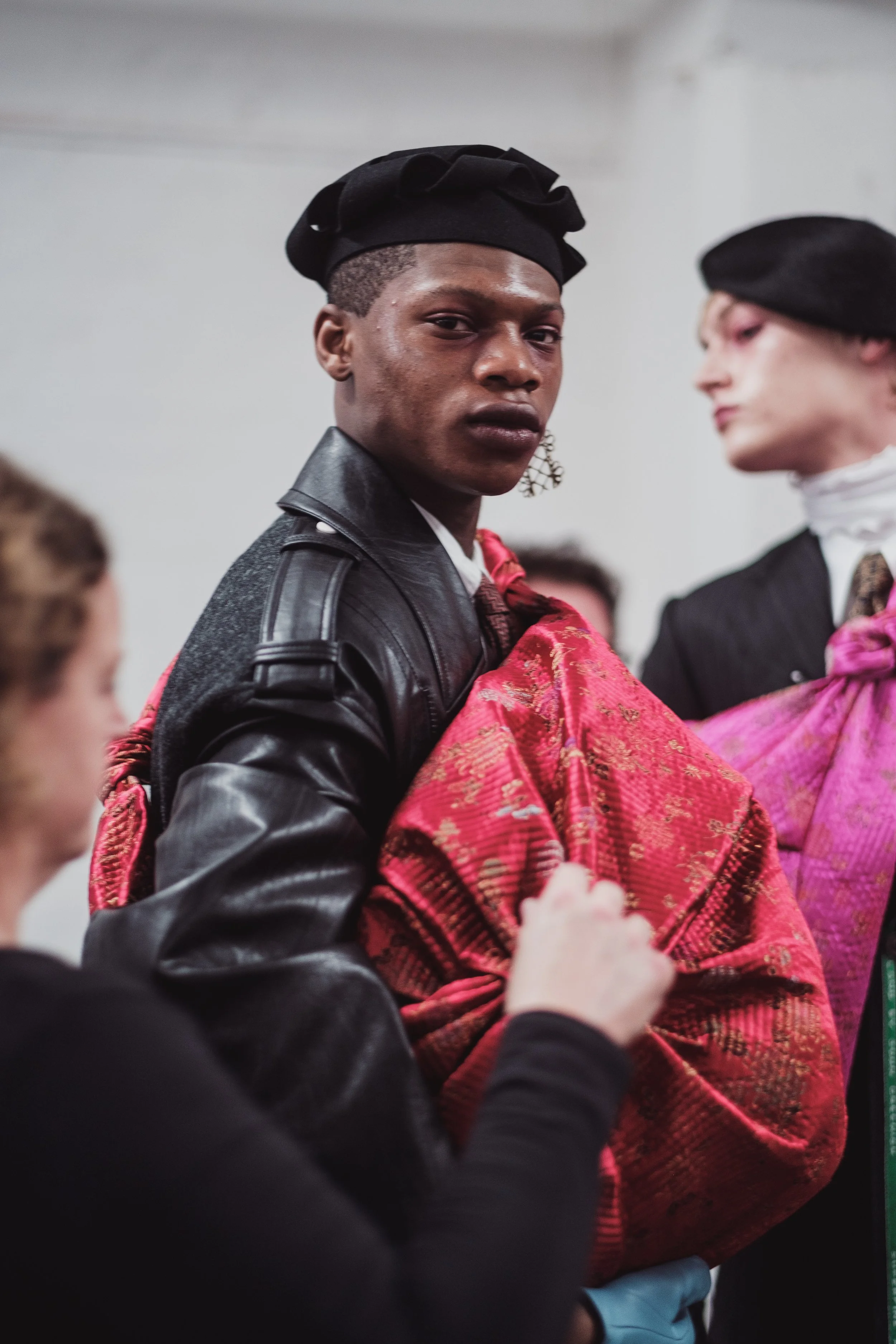 A person wearing a black leather jacket and a black hat, holding a pink and gold fabric, in a room with a white background, participating in what appears to be a fashion event or photo shoot.