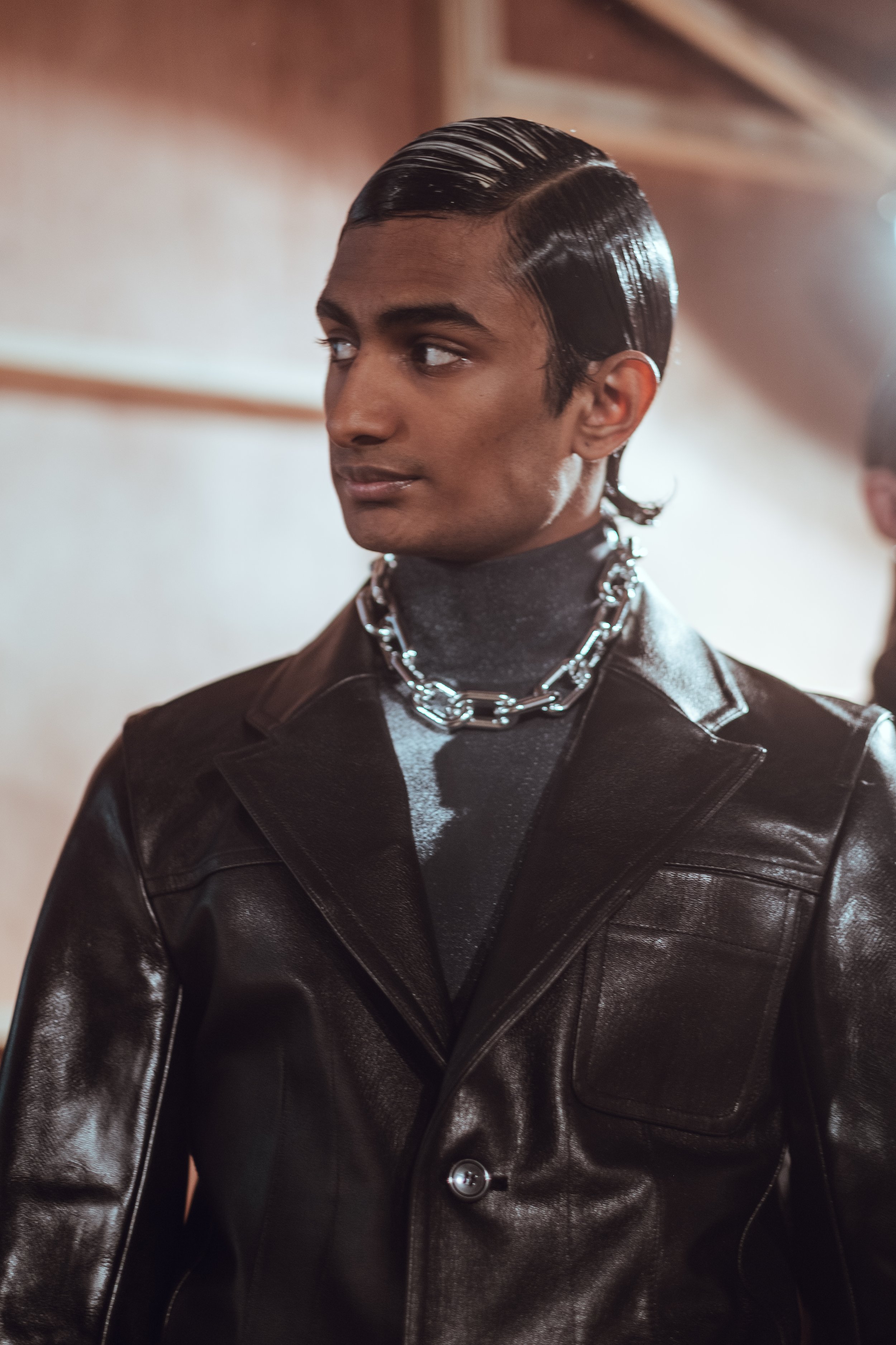 A young man with slicked-back black hair styled in a vintage look, wearing a shiny black leather jacket over a dark turtleneck, accessorized with a large silver chain necklace, standing indoors with soft lighting.