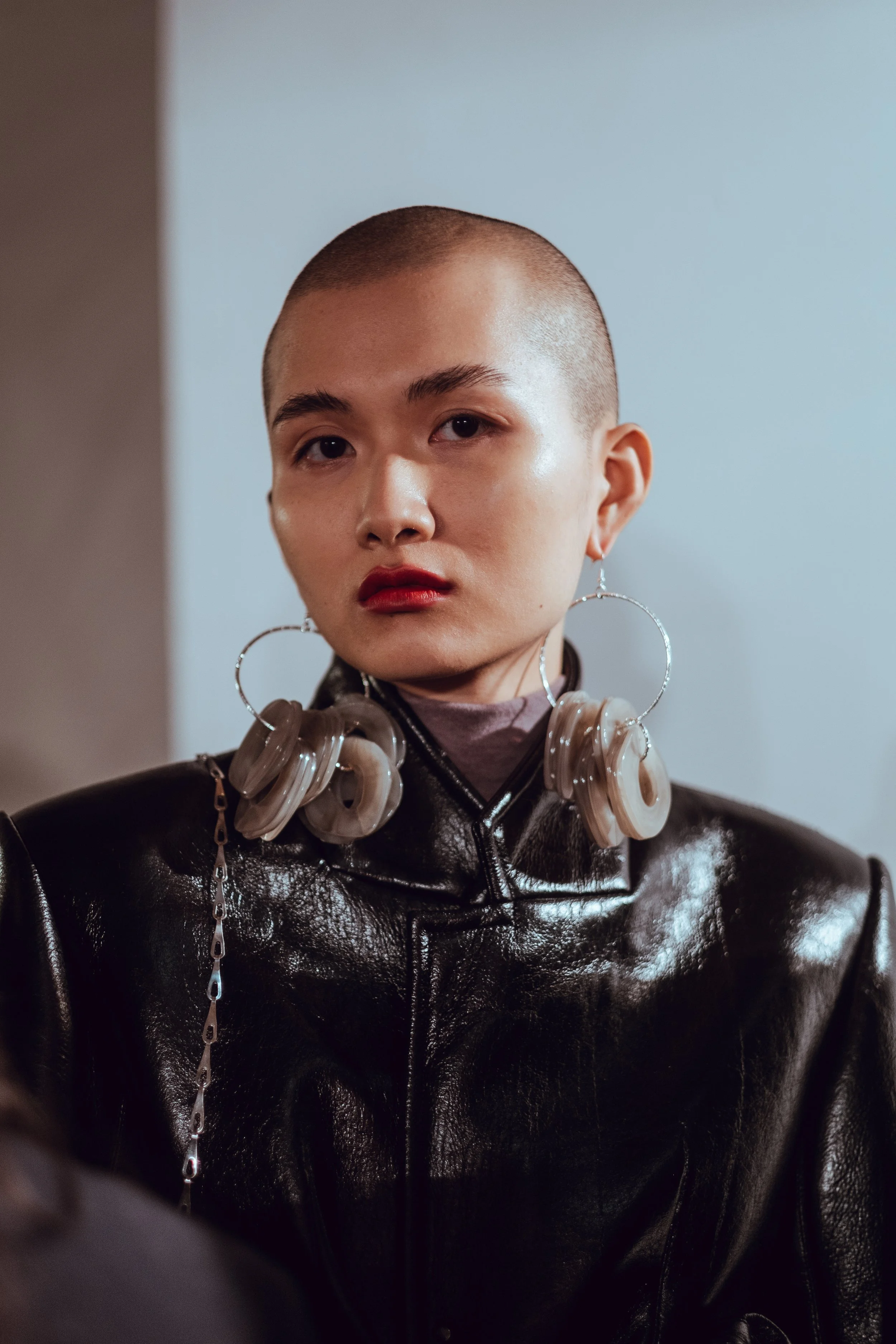 A person with a shaved head wearing large hoop earrings, a dark shiny leather jacket, and makeup, looking directly at the camera with a neutral expression.