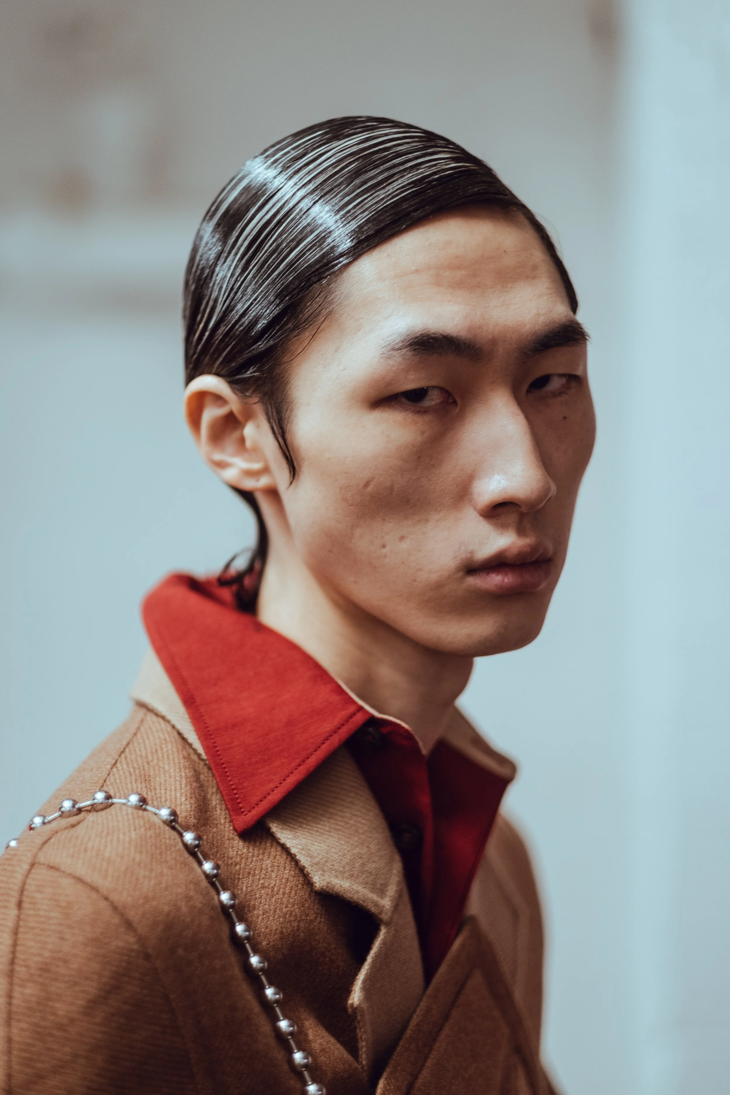 Close-up of a young man with slicked-back dark hair, wearing a brown coat with a red shirt underneath, looking at the camera with a serious expression.