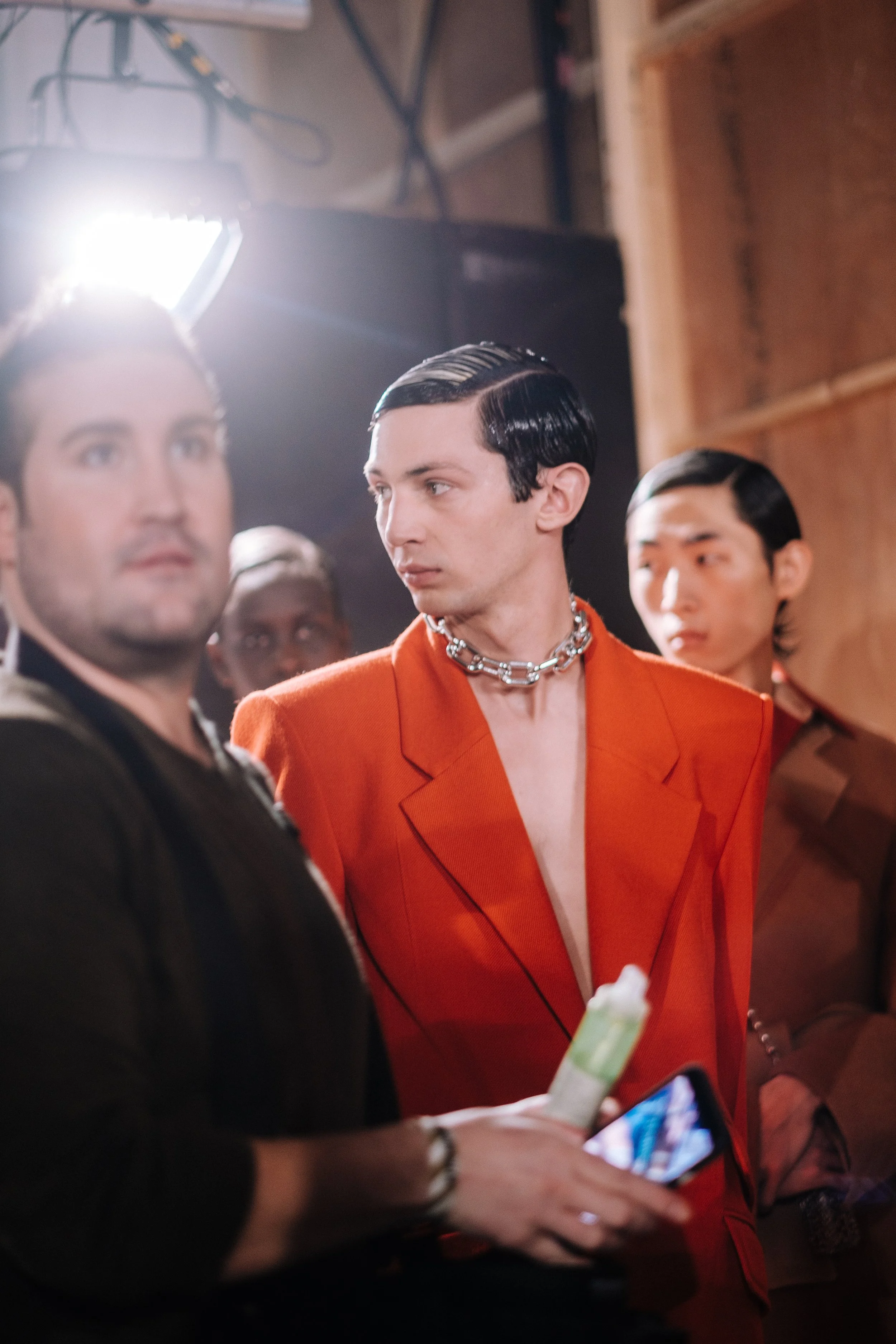 A group of stylish men, one in an orange blazer and a chain necklace, standing together in a backstage setting.