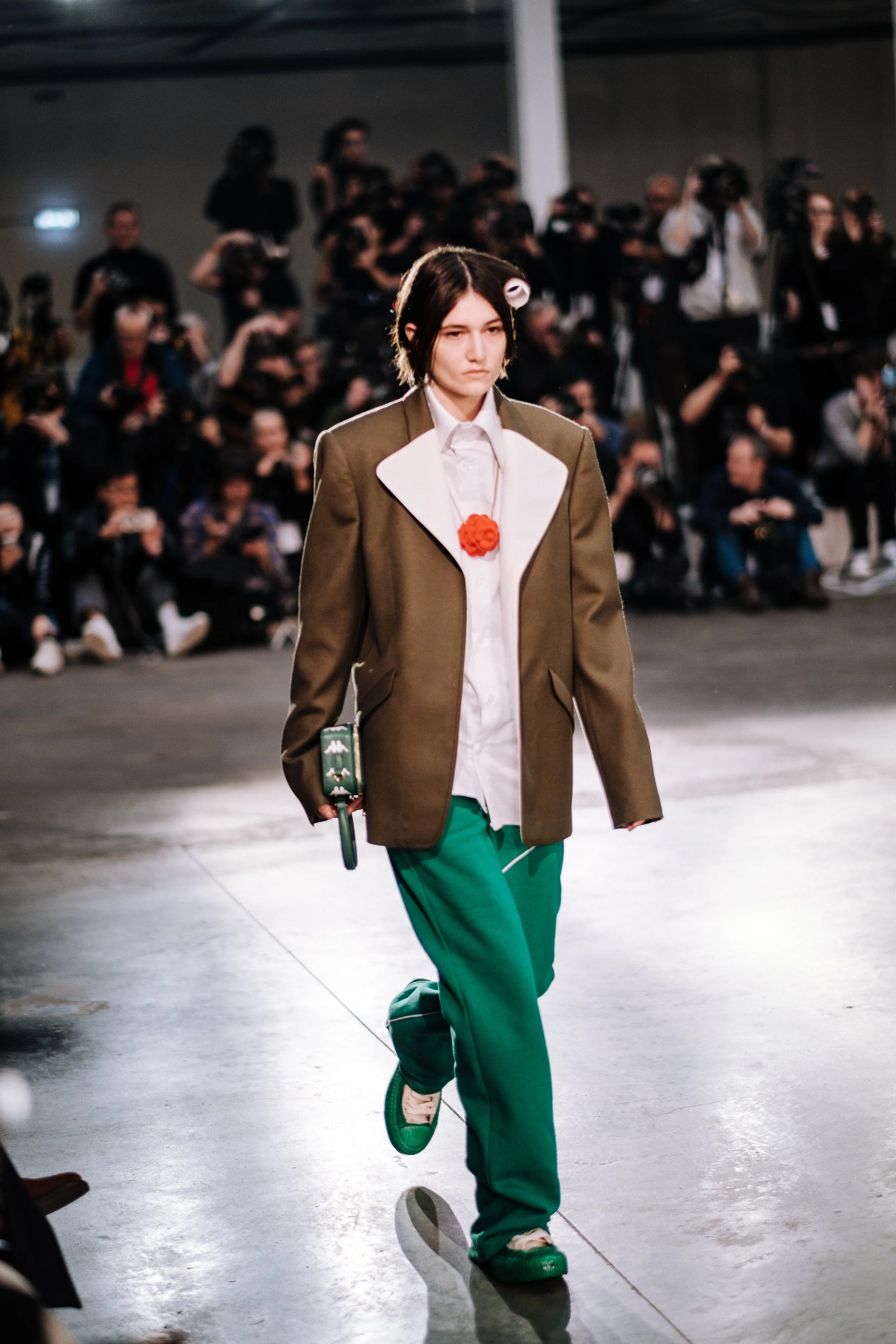 Model walking down the runway at a fashion show wearing an oversized brown blazer over a white shirt, green pants, and green sneakers, with an audience and photographers in the background.