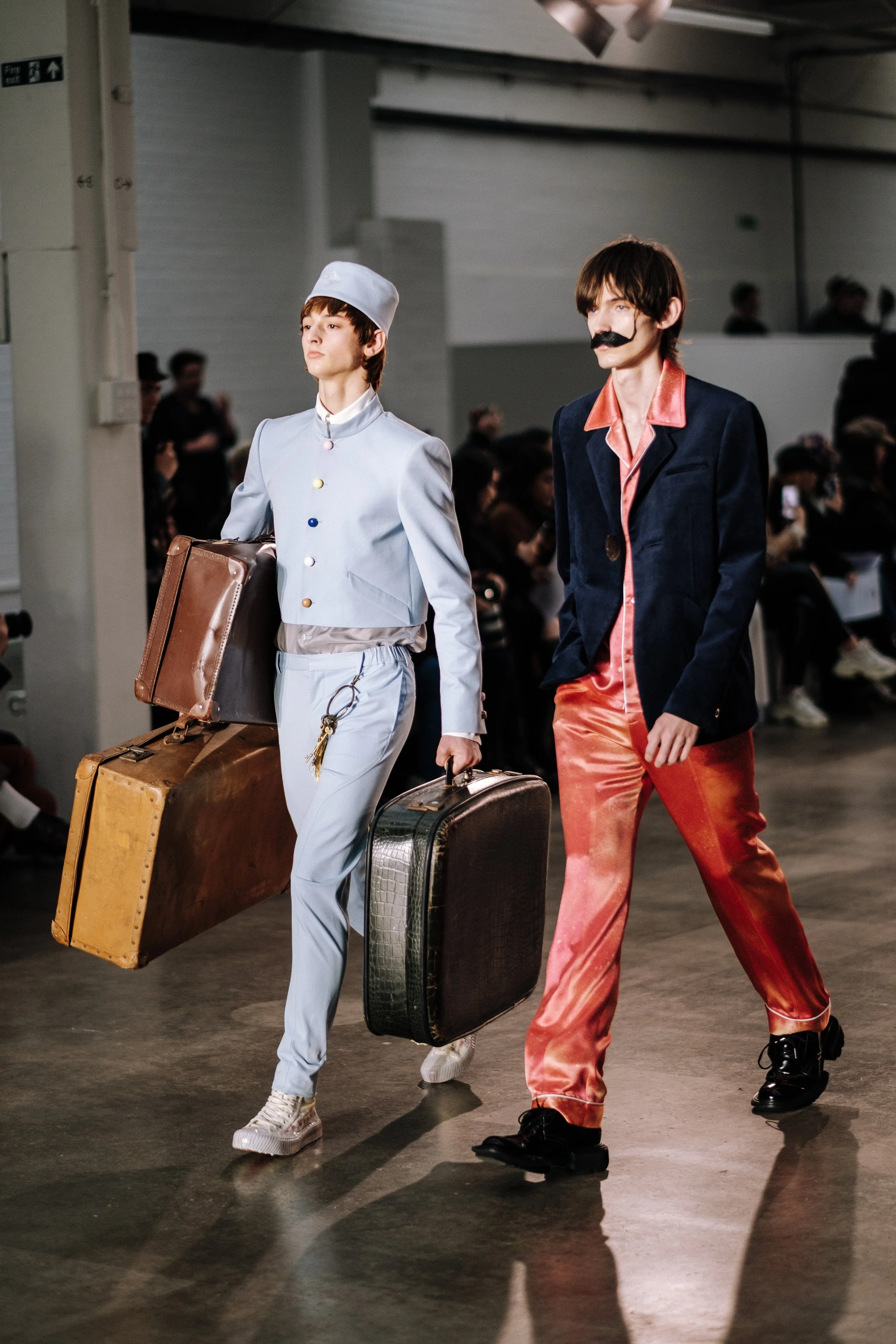 Two male models walking on a runway with vintage luggage, wearing retro-style outfits, in a fashion show setting.