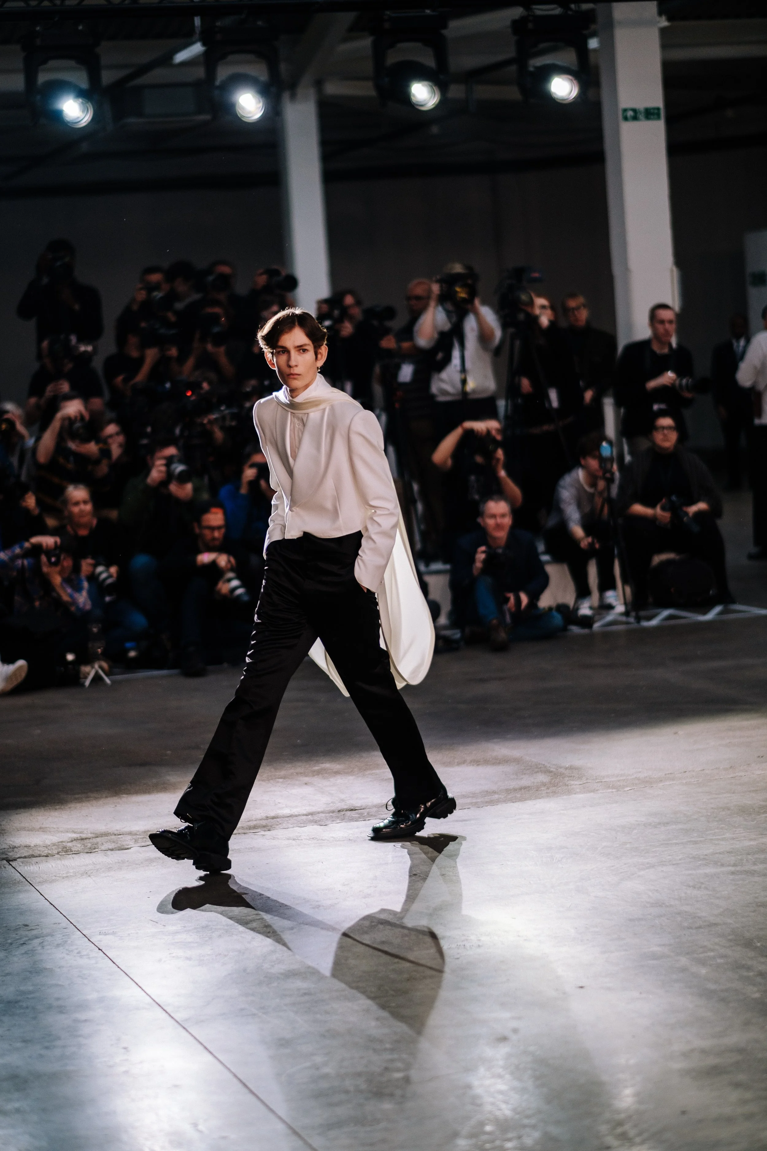 Young male model walking on a runway during a fashion show with photographers and audience in the background.