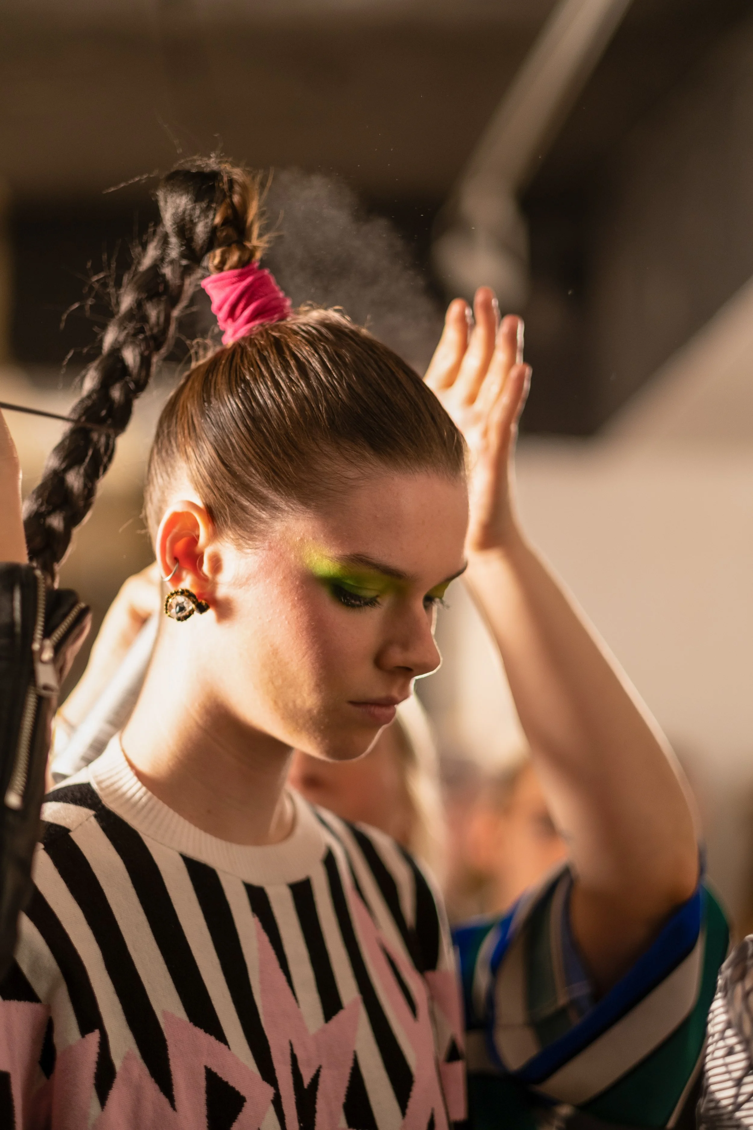 A young woman with her hair in a braided ponytail, wearing a striped shirt with pink accents, and green eyeshadow, is standing with her eyes closed and head bowed in a backstage or dressing room area.