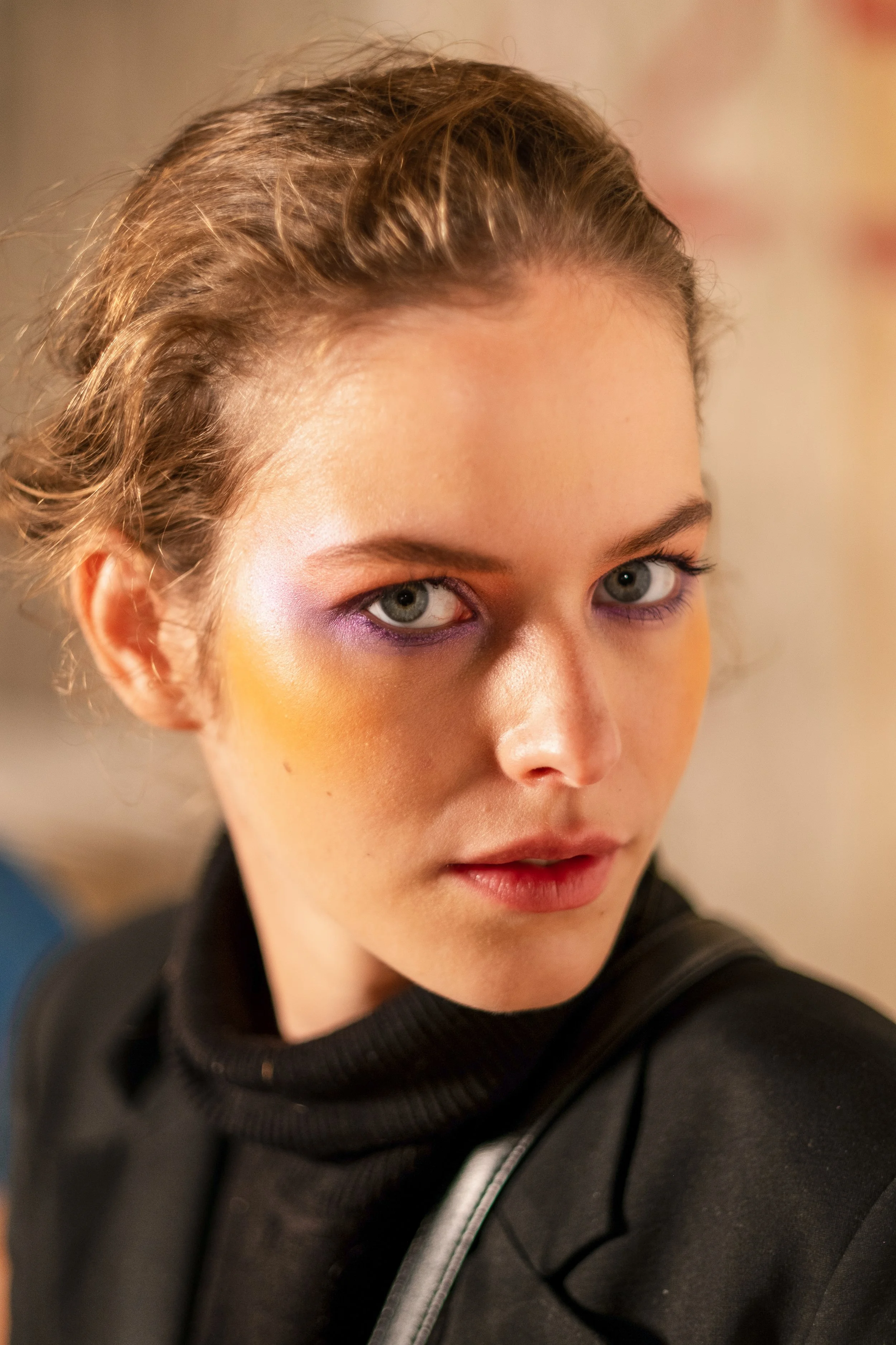 A young woman with light skin, short brown hair, and colorful makeup including purple eyeshadow and orange blush, wearing a black turtleneck and looking over her shoulder.