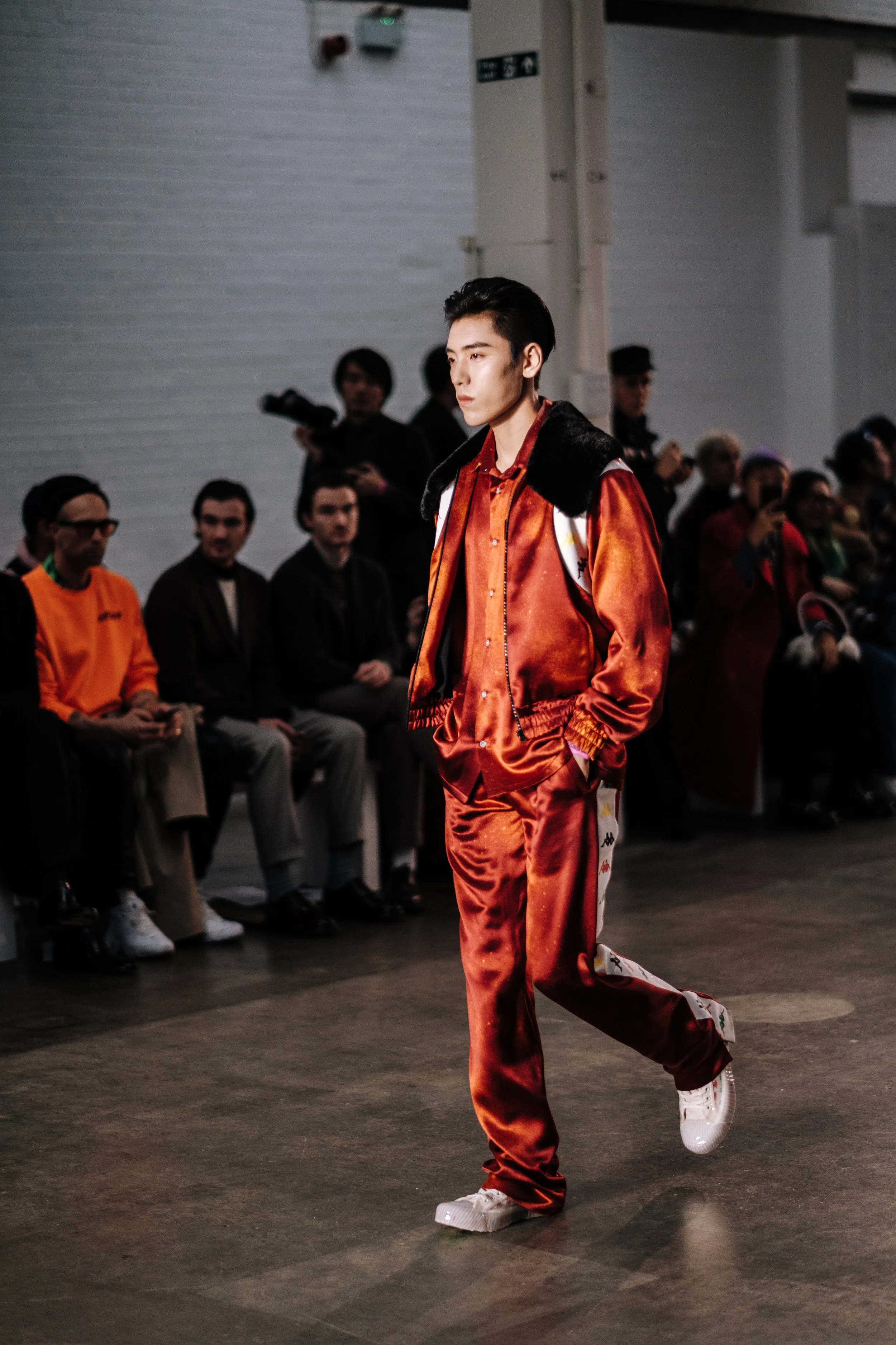 Male model walking on runway at a fashion show, wearing a shiny red tracksuit with white and black details, white sneakers, and a backpack, with seated audience watching in the background.