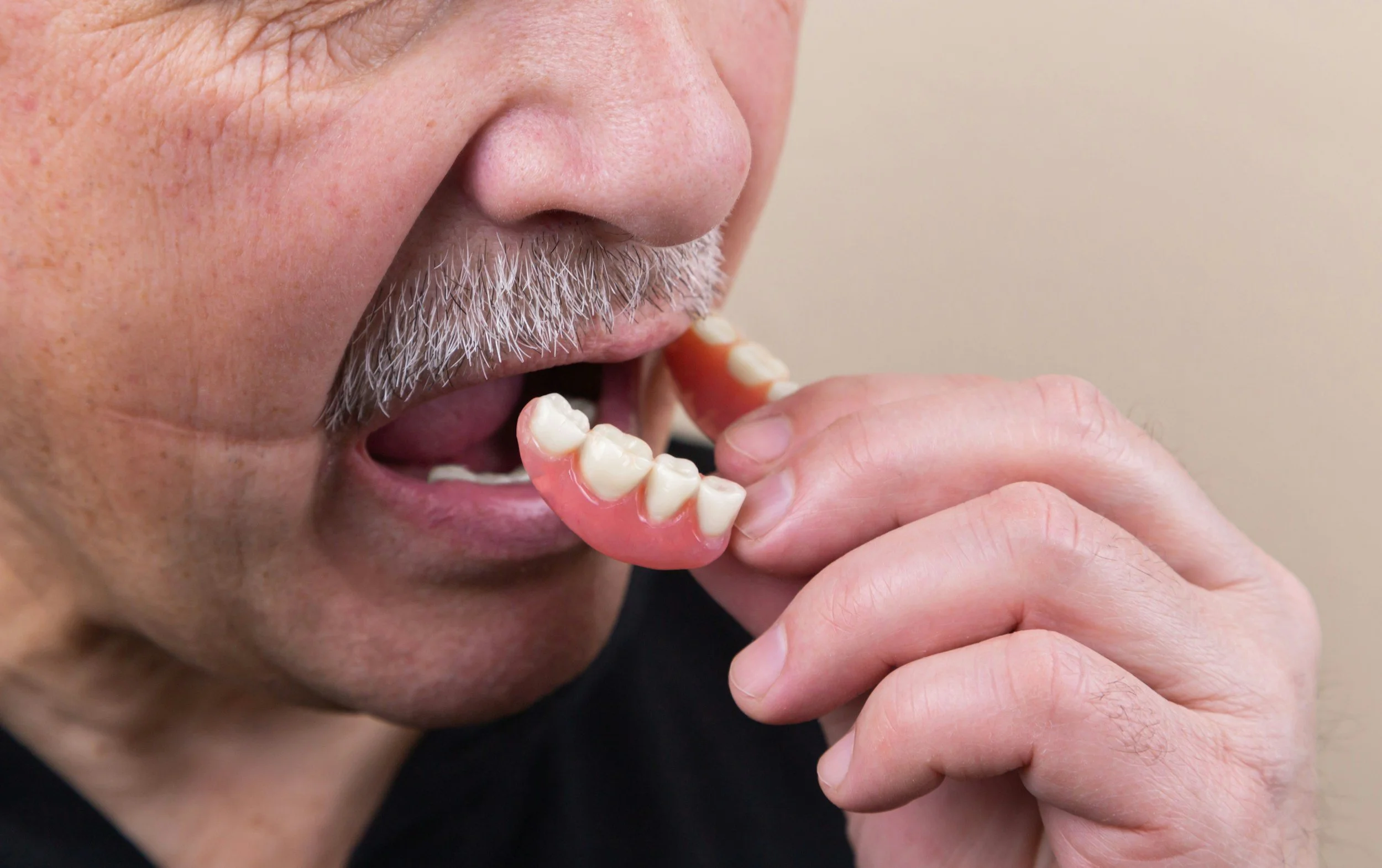A person with gray hair and a mustache is holding a denture near his mouth.