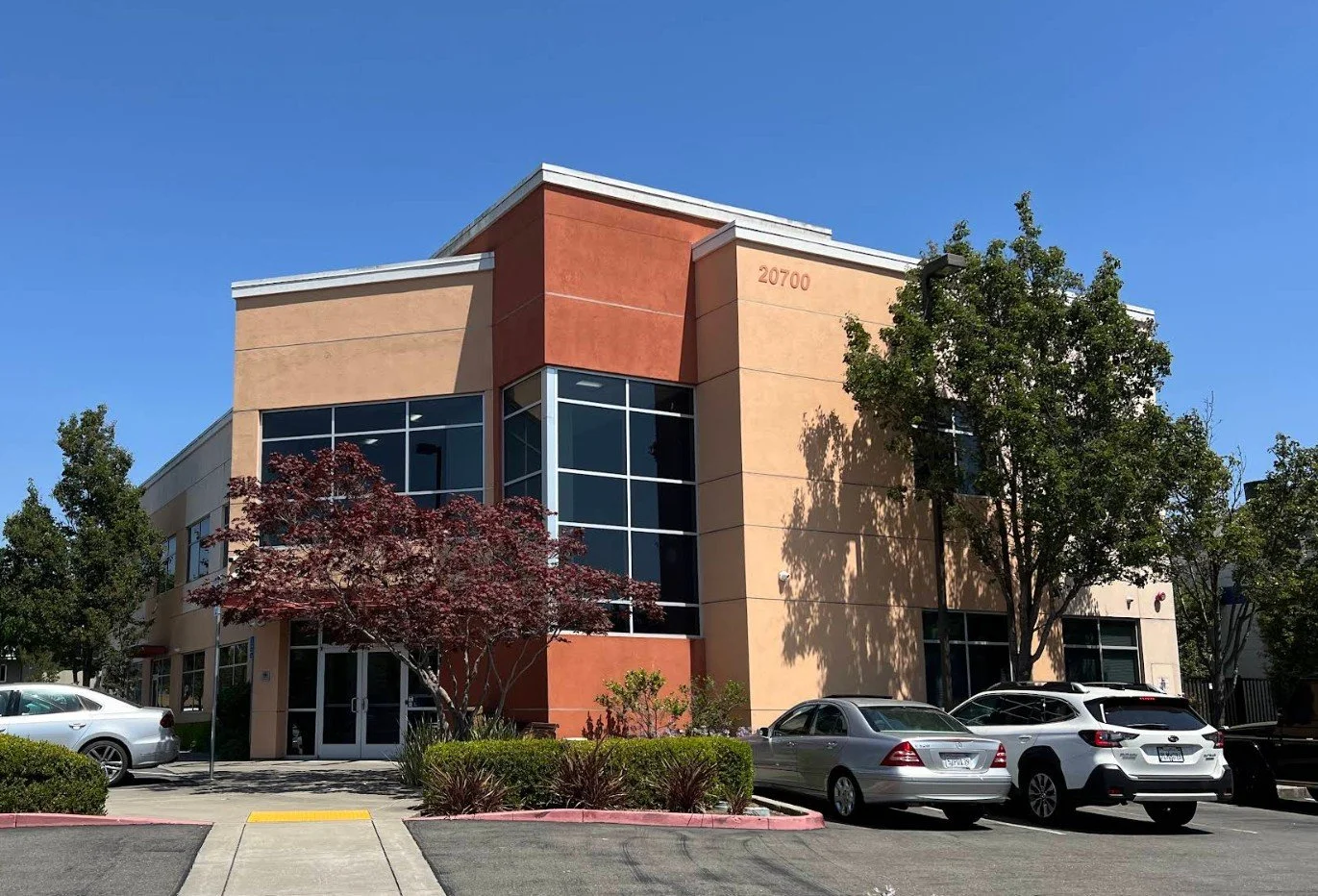 Modern two-story commercial building with beige and reddish-orange exterior, large glass windows, and a parking lot with several cars in front. There are trees and bushes around the entrance and a clear blue sky overhead.