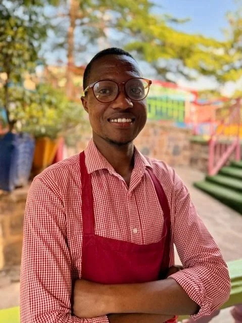 A man wearing glasses and a red and white checkered shirt with a red apron, standing outdoors with trees, colorful structures, and stairs in the background.