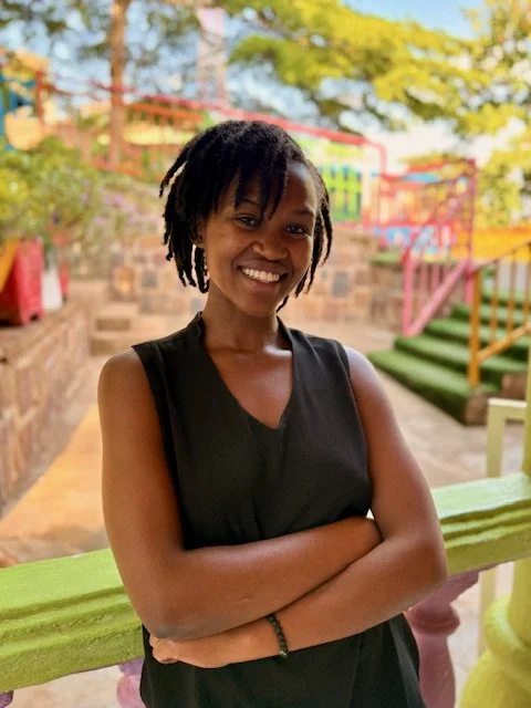 A smiling woman with shoulder-length dreadlocks wearing a sleeveless black top, standing outdoors in front of colorful stairs and a stone wall, with trees and sky in the background.