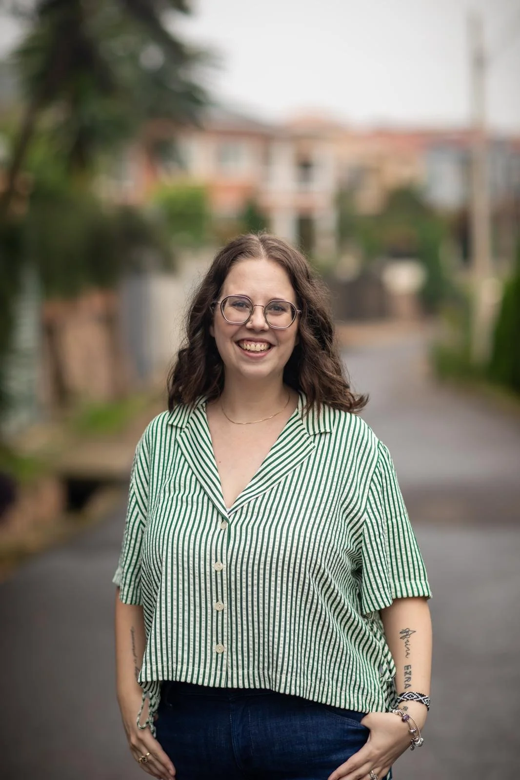 A young woman with glasses smiling outdoors on a street, wearing a green and white striped shirt and dark pants.