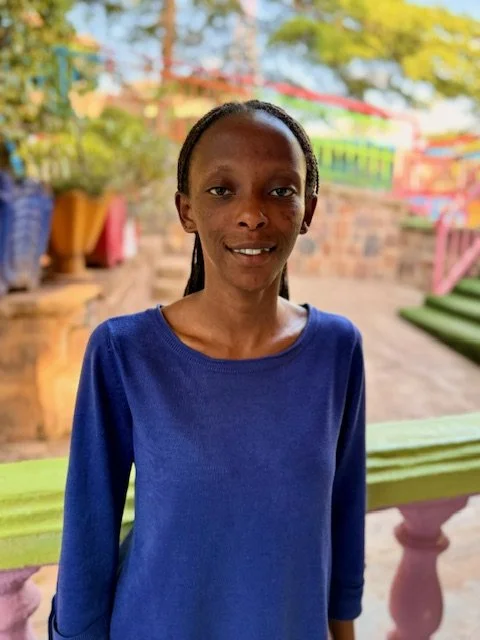 A young woman with dark skin and long black hair in a ponytail, wearing a blue long-sleeve shirt, standing outdoors with colorful trees and structures in the background.