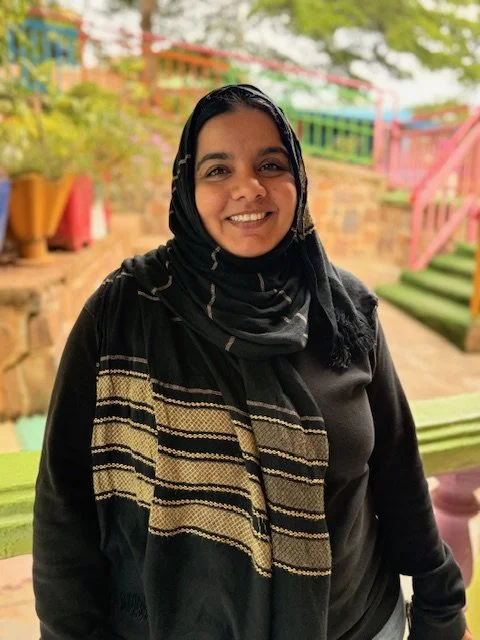 Woman with a black and beige striped headscarf smiling outdoors in a colorful garden setting with steps and greenery in the background.