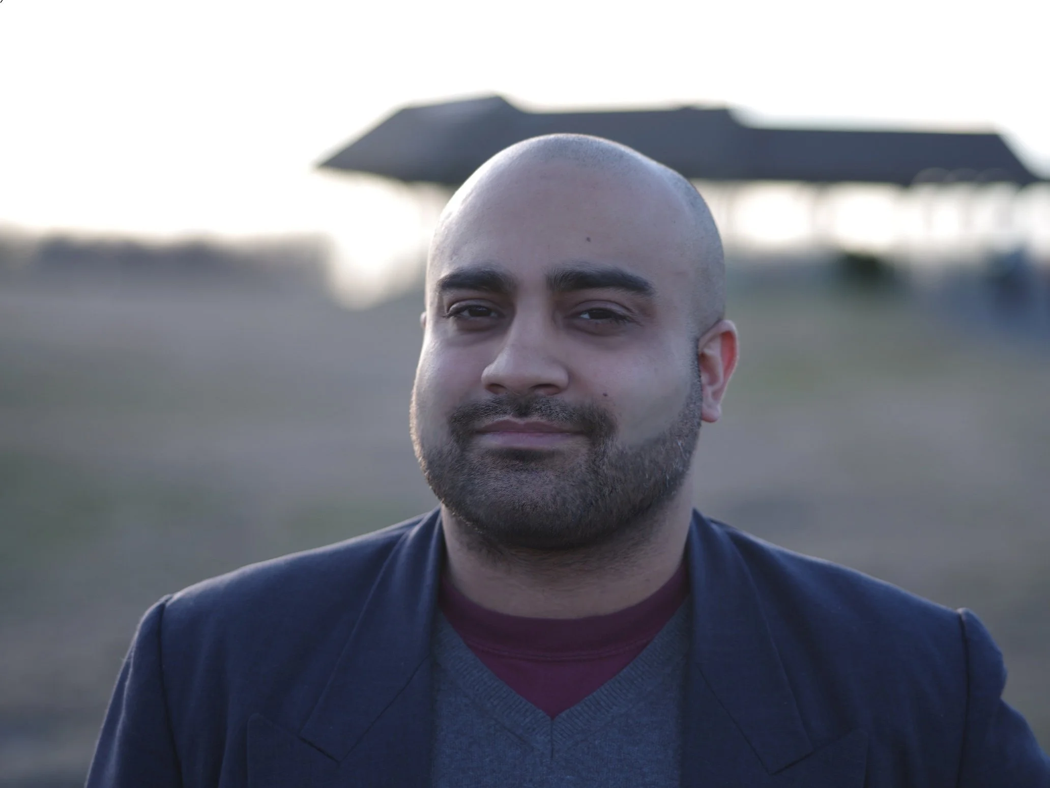 A man with a bald head and beard wearing a maroon undershirt with a gray sweater and a dark blazer, standing outdoors with a blurred background.