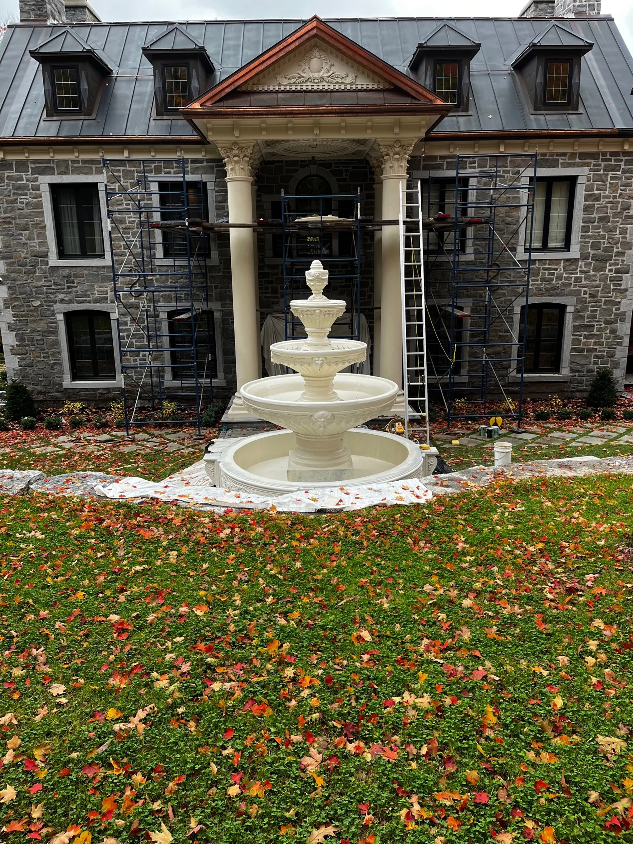 A stone building with a metal roof features a classical facade with columns and a three-tiered fountain in front. The facade is under renovation, with scaffolding and ladders present. The grass lawn is covered with autumn leaves.