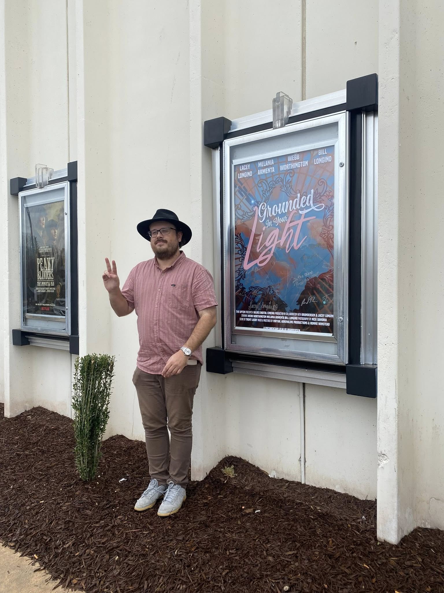 A man with glasses, a beard, wearing a black hat, pink shirt, and gray pants, making a peace sign with his right hand, standing in front of movie posters outside a theater.