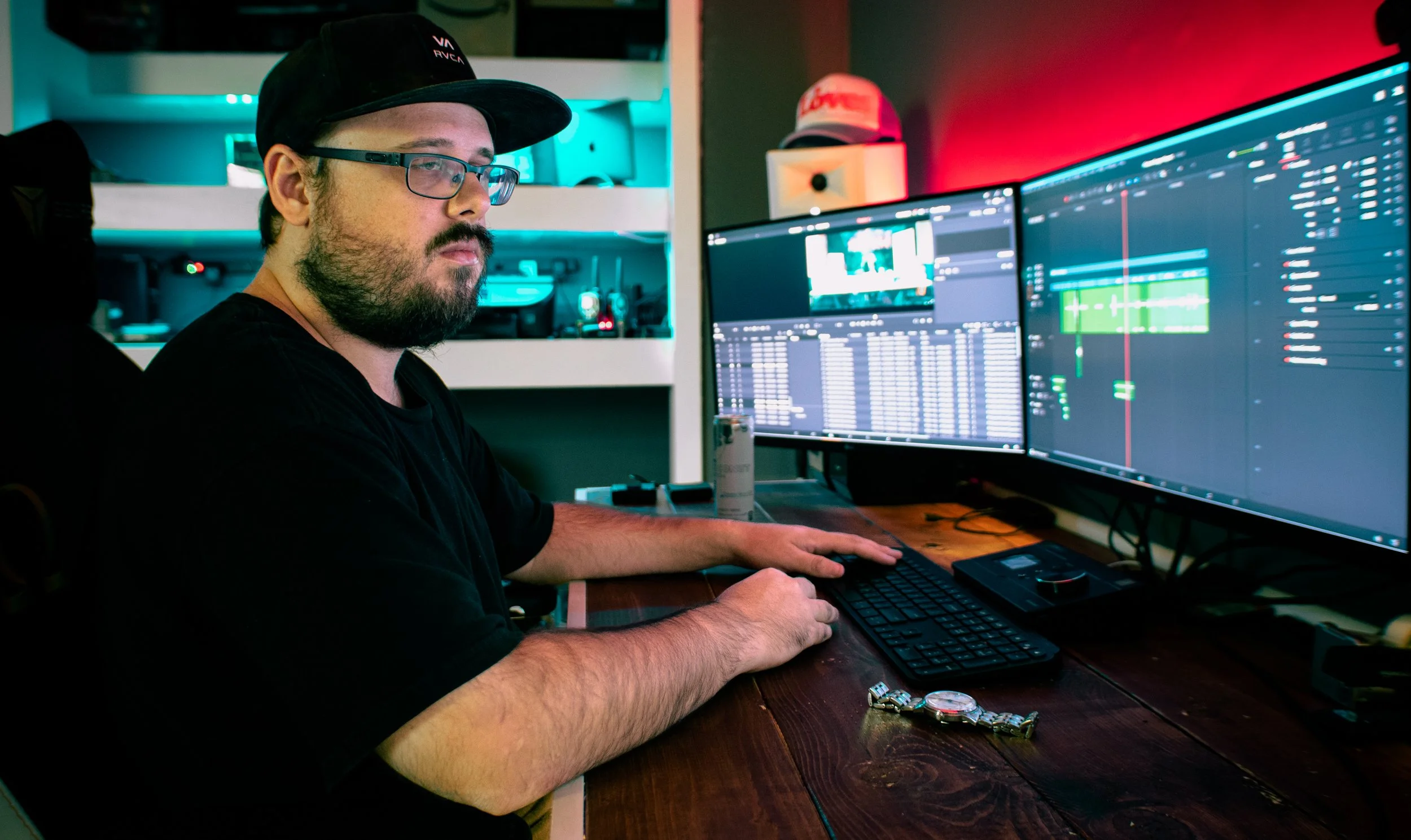 A man with a beard and glasses working at a computer with dual monitors showing audio editing software in a dimly lit room with colorful backlighting.