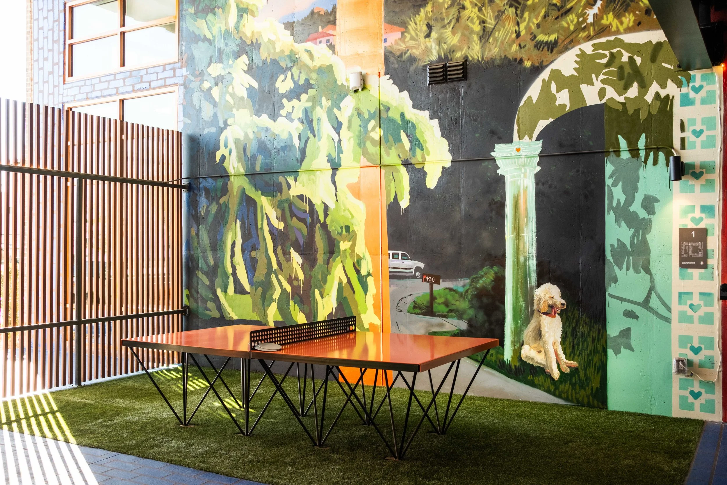 Interior view of a room with a colorful mural of a map and greenery on the wall. There is a wooden table with a black metal frame and two chairs. The floor is covered with green artificial grass, and sunlight creates striped shadows on the floor.