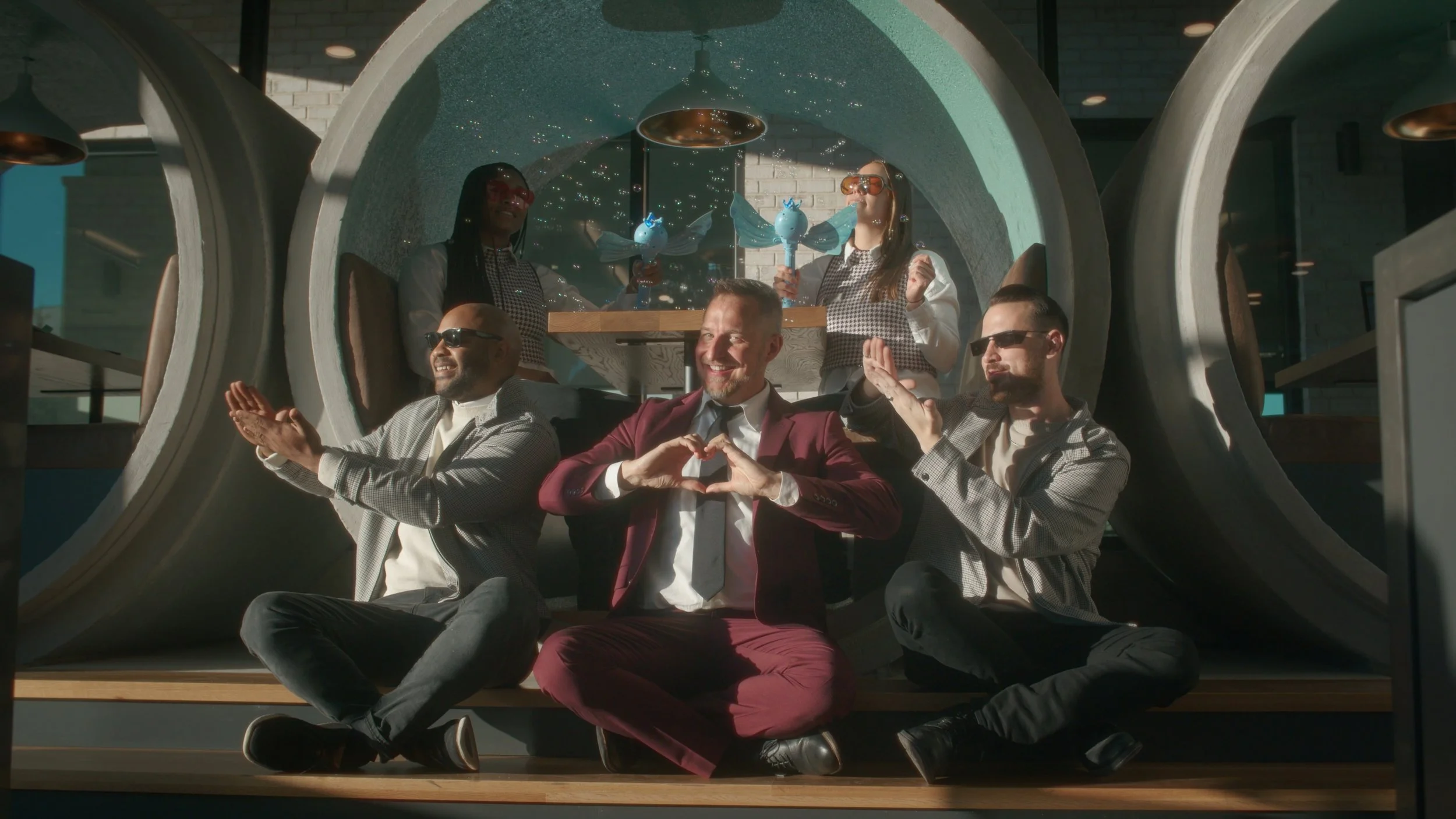 A man in a burgundy suit making a heart shape with his hands, flanked by two men in checkered jackets, and surrounded by two women dressed as fairy tale characters with wings and masks, inside a futuristic circular structure with sunlight streaming in.
