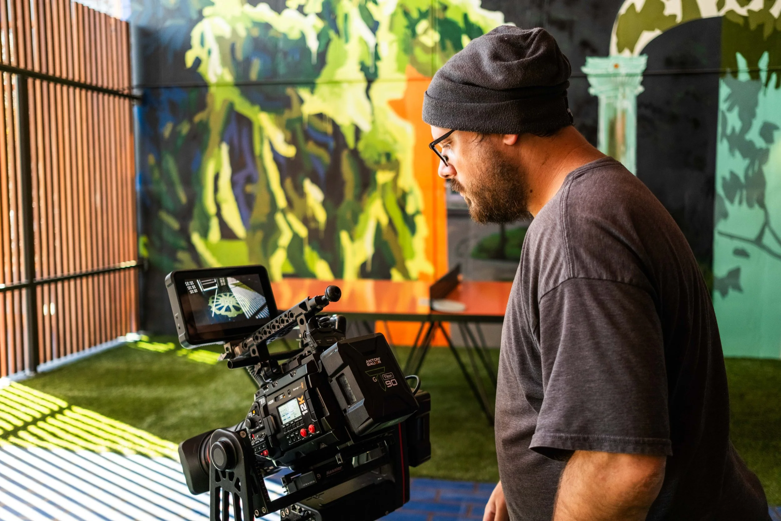 A man with a black beanie, glasses, and a gray shirt operating professional video camera equipment on a set with colorful, abstract background paintings.