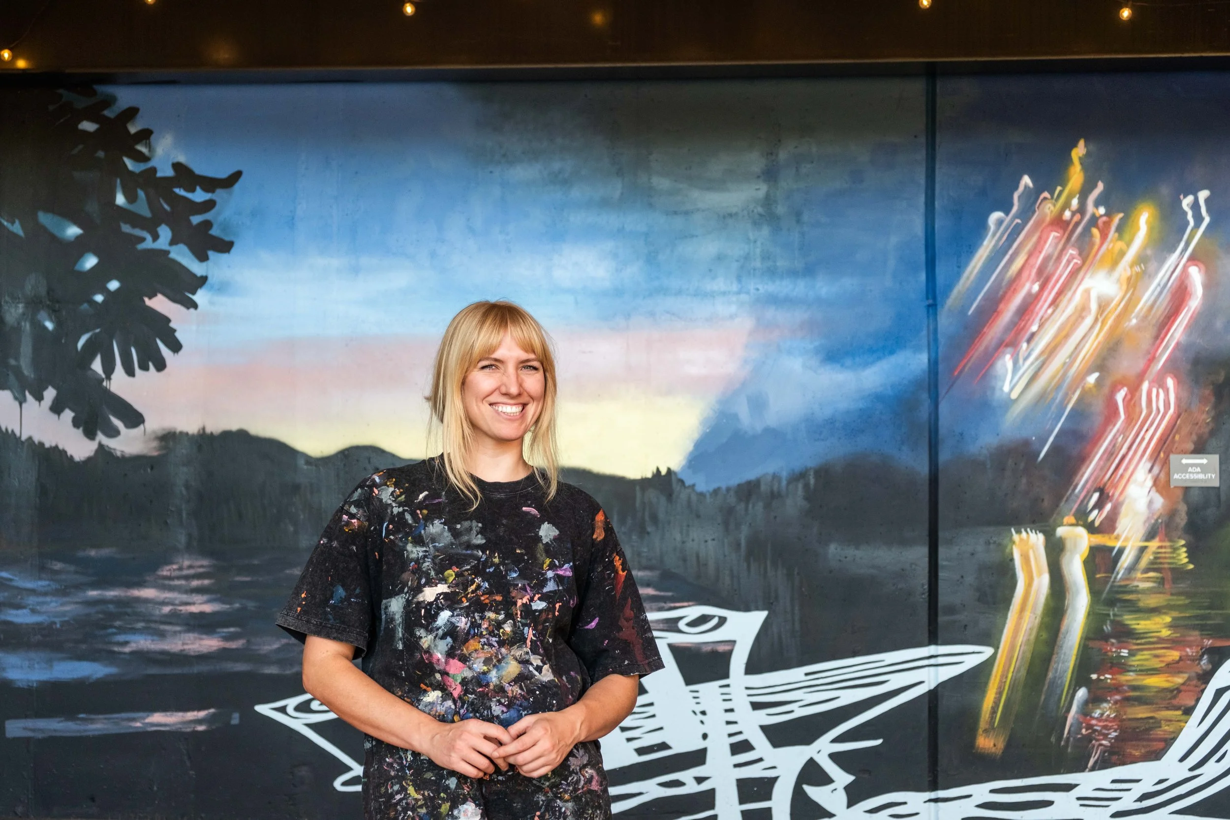 Young woman with blonde hair, wearing a black t-shirt splattered with multicolored paint, smiling and standing in front of a painted mural background with a tree silhouette, a sunset, and abstract colorful lines.